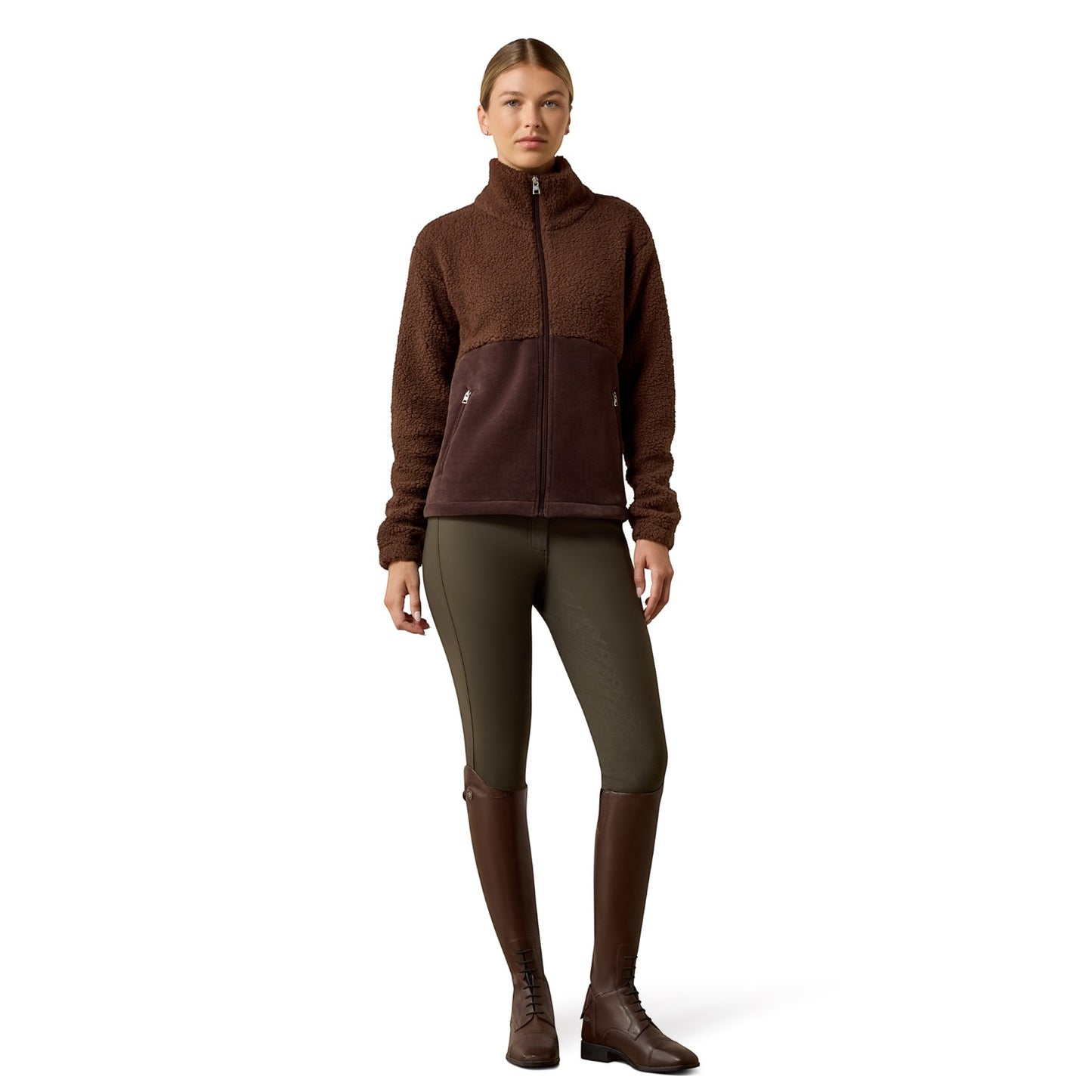 A woman modeling a brown fleece zip-up jacket with dark brown and black color blocking, paired with olive green riding tights and dark brown riding boots, against a plain white background.