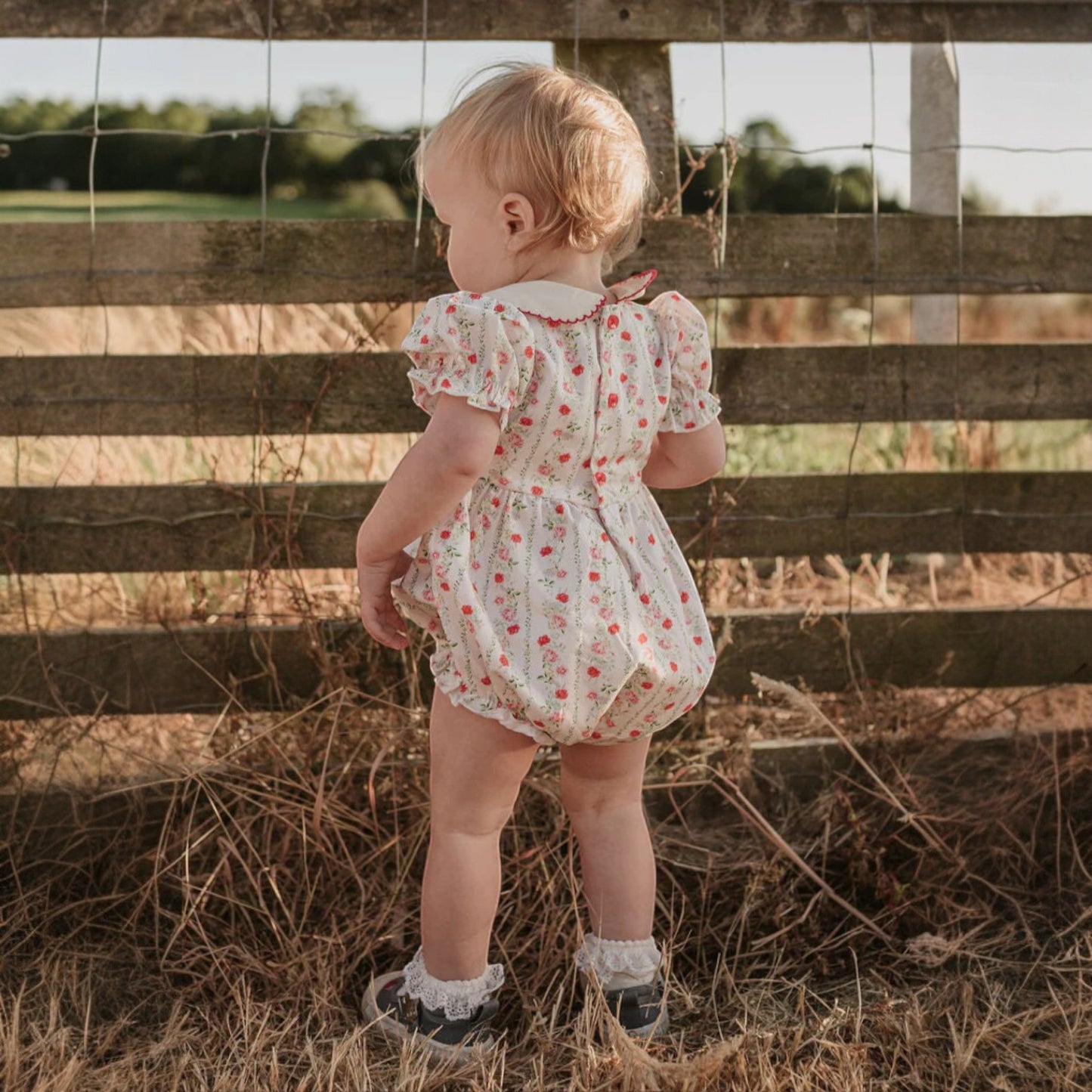 Child in a floral dress standing next to a wooden fence in a field.
