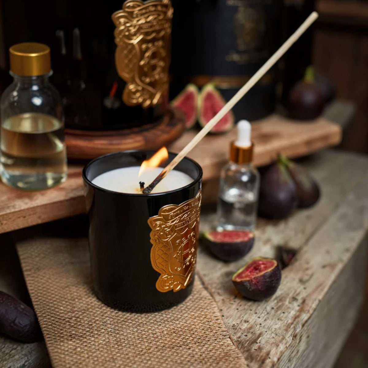 A black scented candle with a gold decorative emblem and a lit wick, styled on a rustic wooden surface with figs, small glass bottles, and a matchstick in the foreground.