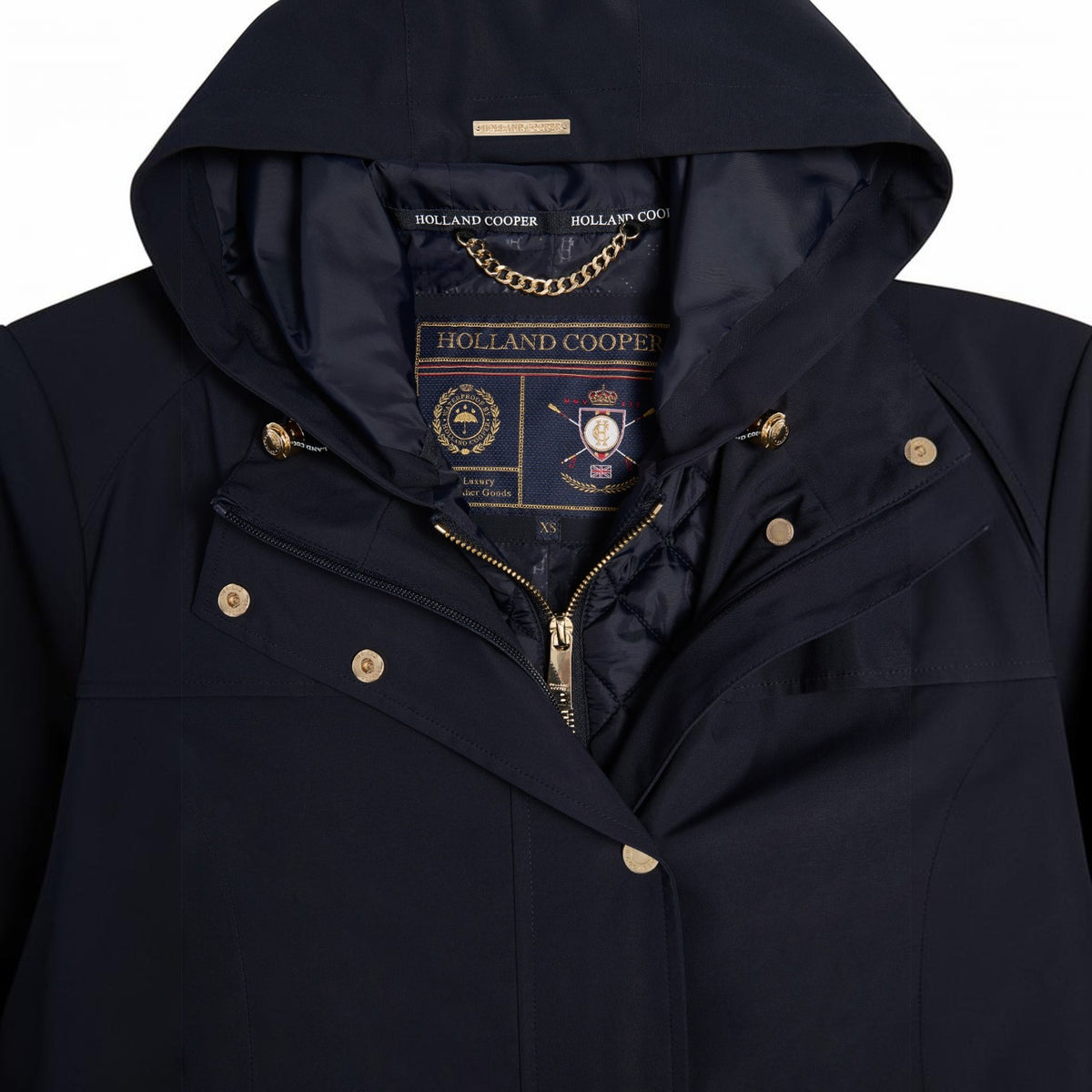 Close-up of a navy blue hooded jacket by Holland Cooper, showing the hood lining, gold-tone zipper, snap buttons, and the brand label inside the collar.