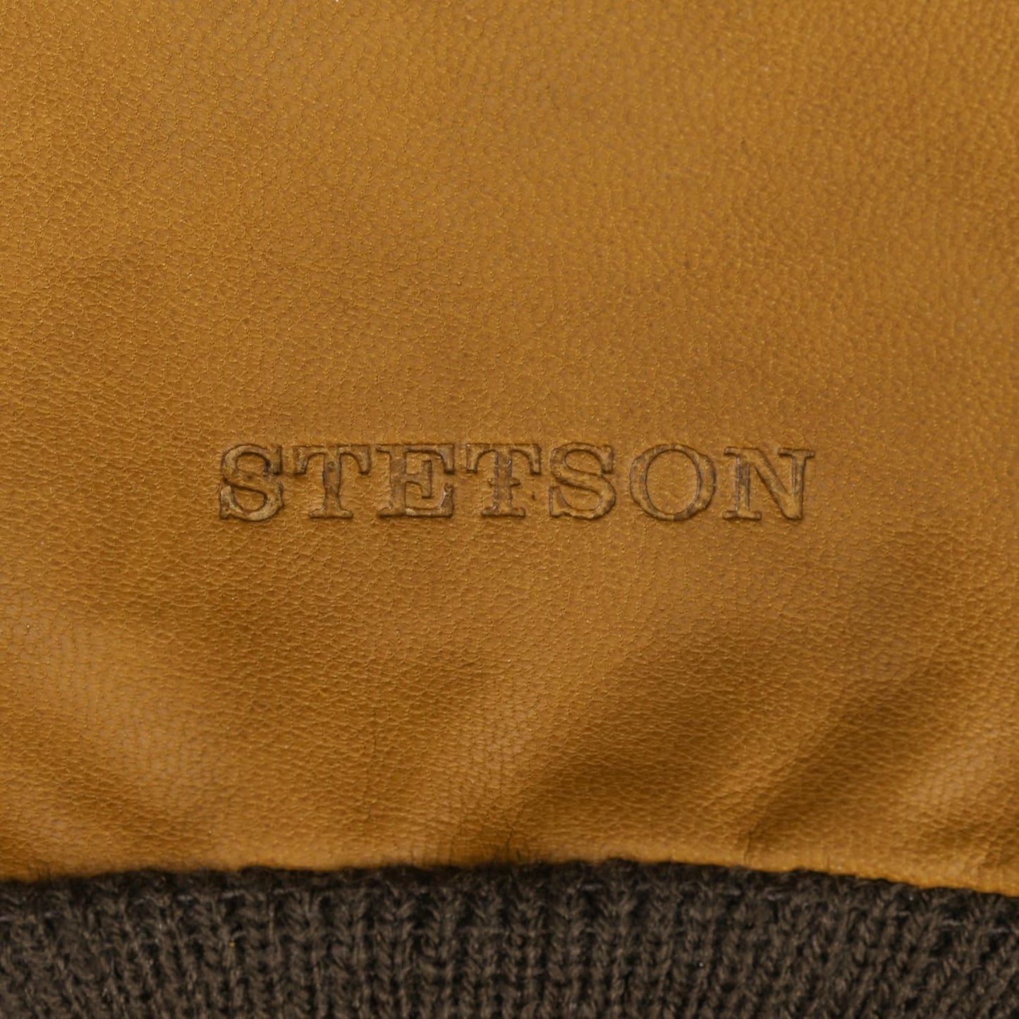 Close-up of a tan leather product with the embossed brand name 'STETSON' and a dark ribbed knit trim at the bottom.