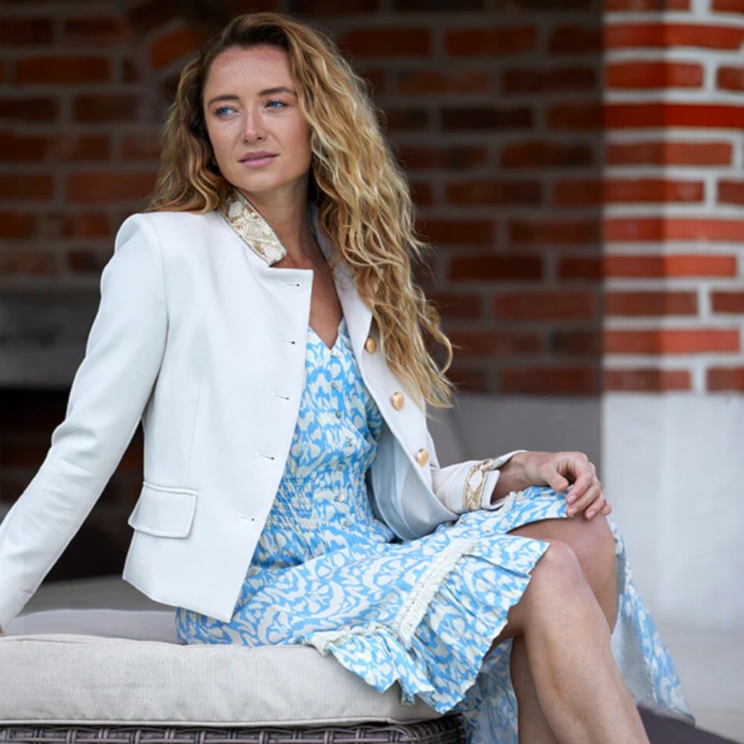 Woman in a white blazer and blue dress sitting against a brick wall.