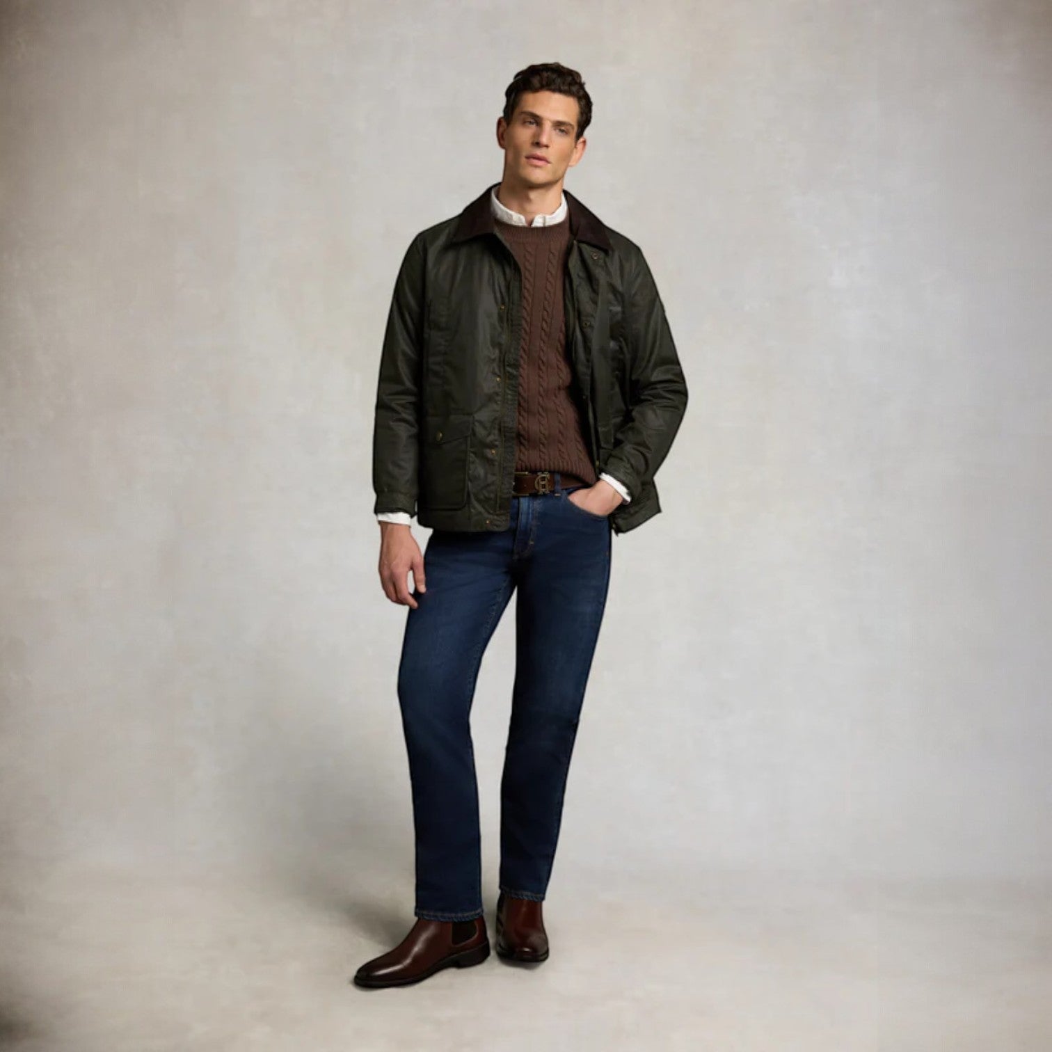 Man wearing a dark green jacket, brown shirt, blue jeans, and brown shoes on a plain background