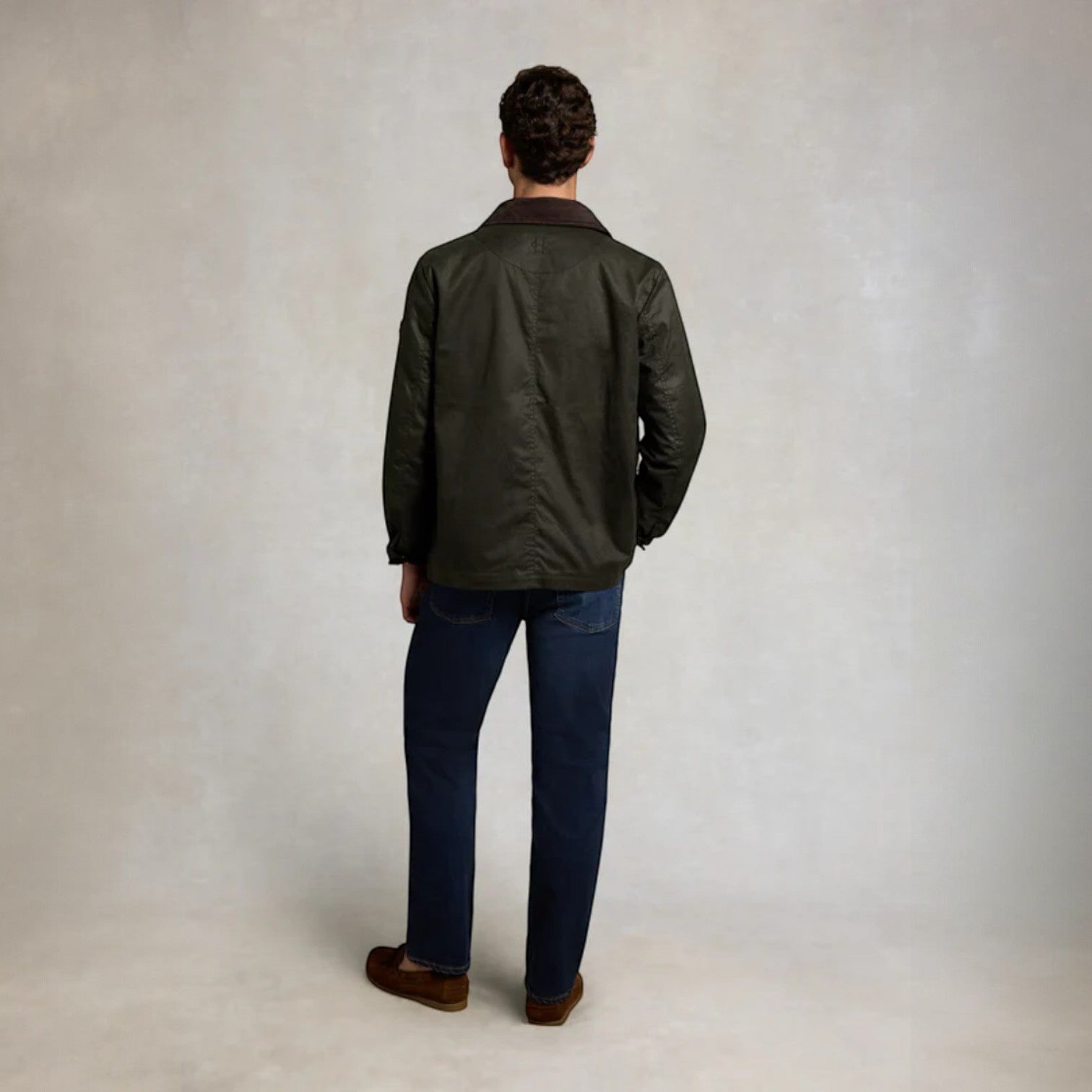 Man wearing a dark green jacket and blue jeans standing against a plain background
