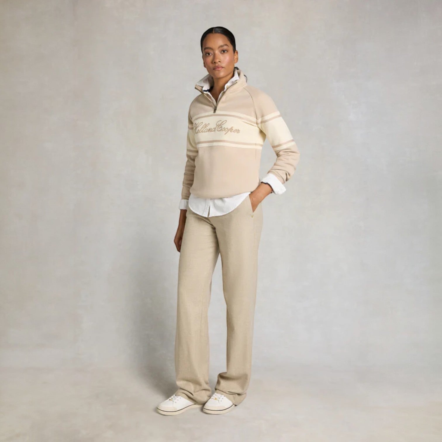 Person wearing a beige sweater with a logo and light-colored pants on a plain background