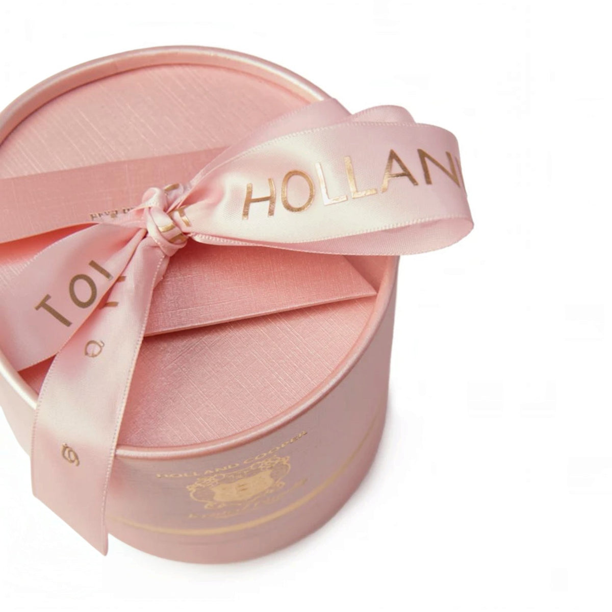 A round pink gift box with a matching satin ribbon tied in a bow. The ribbon is light pink with gold foil lettering that reads 'TO THE HOLLAND'.