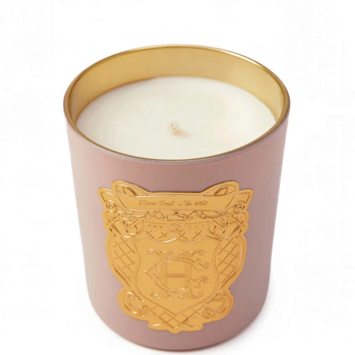 A luxury scented candle in a matte dusty rose pink glass jar with a shiny gold crest emblem on the front and a gold-rimmed interior, filled with white wax.