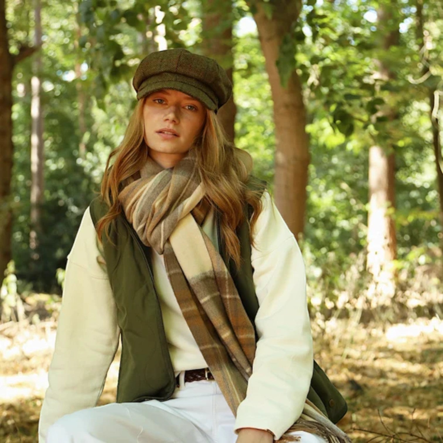 A woman sitting in a sunlit forest wearing a brown tweed newsboy cap, an olive green vest, a cream fleece jacket, white trousers, and a beige-and-brown plaid scarf.