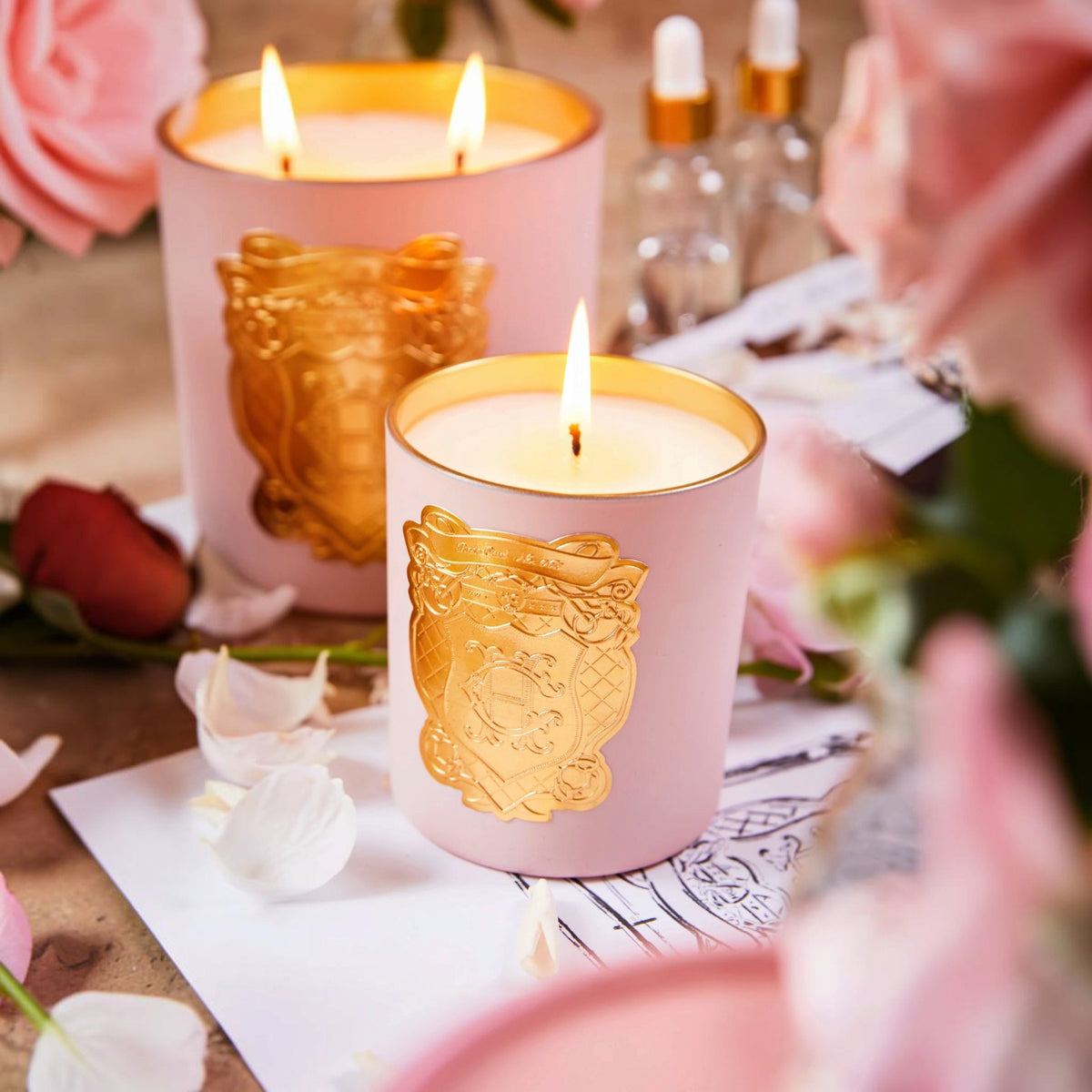 Two lit pink candles with gold trim and embossed gold crest designs, styled with pink roses, rose petals, and small glass dropper bottles on a tabletop.