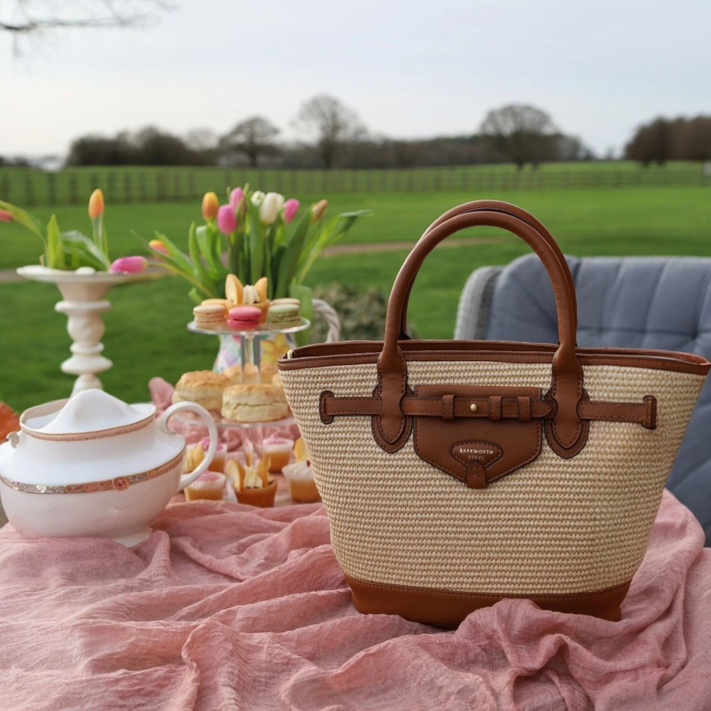 Beige and brown handbag on a pink blanket with a tea set and tulips in the background.