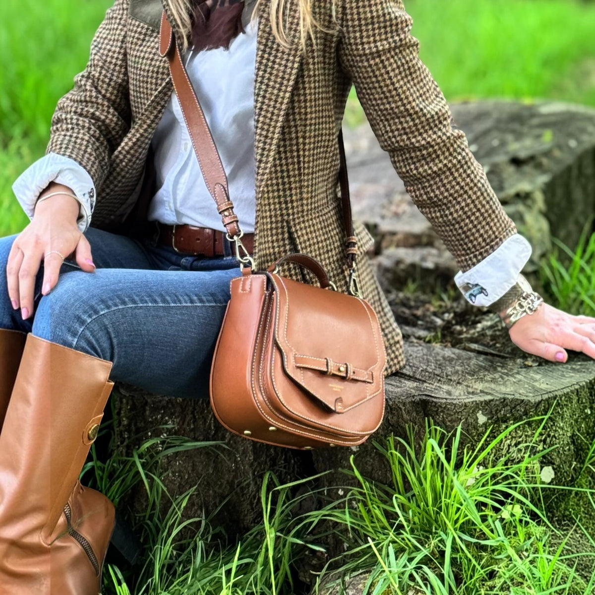 A woman sitting outdoors on a stone wall wearing a brown houndstooth blazer, blue jeans, and tan knee-high boots, with a brown leather crossbody bag resting against her leg.