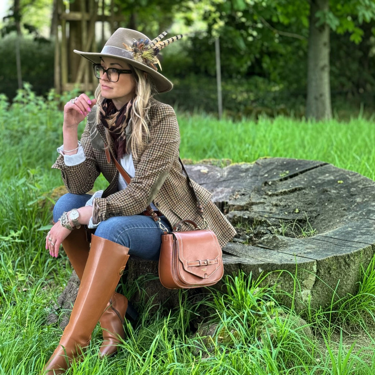A woman sits on a large tree stump in a grassy outdoor setting, wearing a brown houndstooth blazer, matching wide-brim hat with feather accents, blue jeans, tall brown leather boots, and a brown leather crossbody bag.