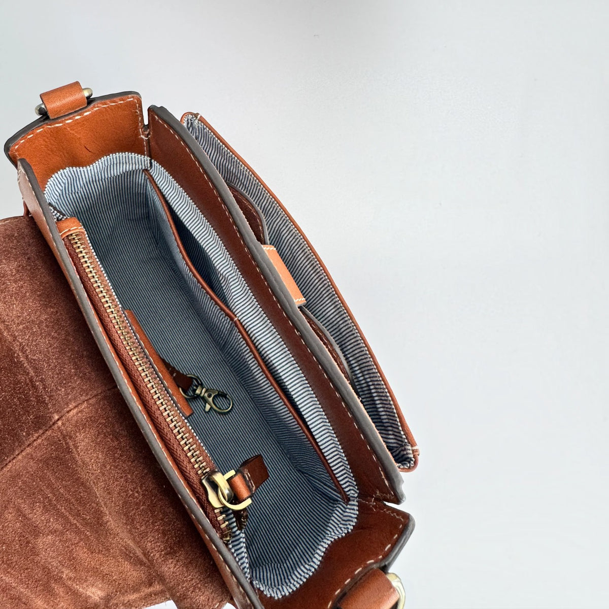 An open brown leather and suede handbag shown from above, revealing a striped blue-gray interior lining with gold-tone zipper hardware and a small pouch inside.