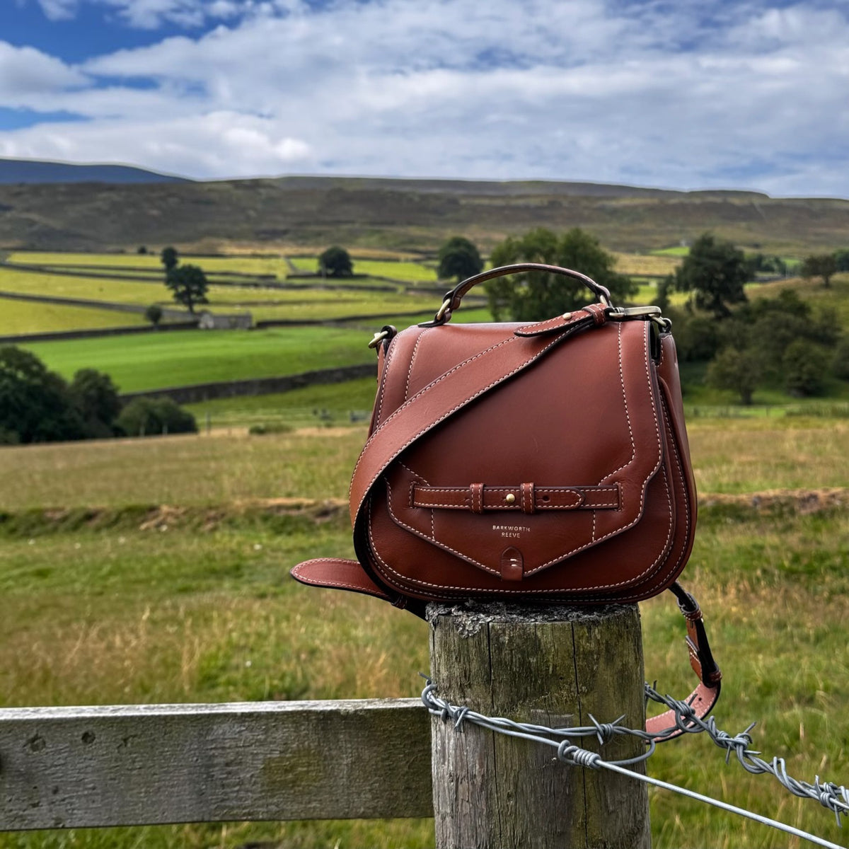 A brown leather shoulder bag with a top handle and shoulder strap, displayed on a wooden fence post in an outdoor rural landscape with green fields and hills under a blue sky.