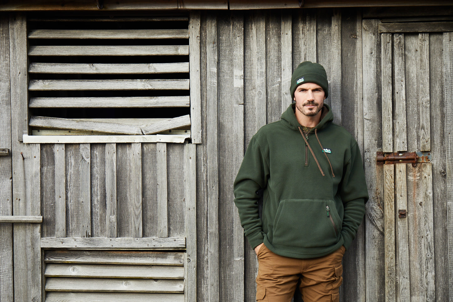 A man wearing an olive green hoodie and matching beanie stands against a weathered gray wooden barn wall, with his hands in his pockets.