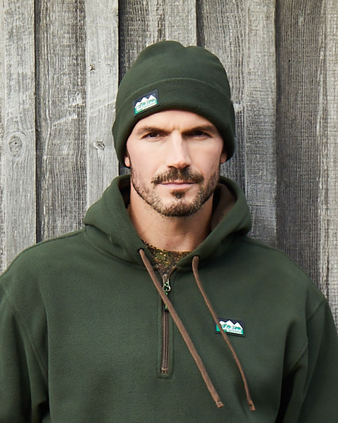 A man wearing a dark green fleece pullover hoodie and matching knit beanie, both featuring a small mountain logo patch on the chest, standing against a rustic gray wooden plank wall.