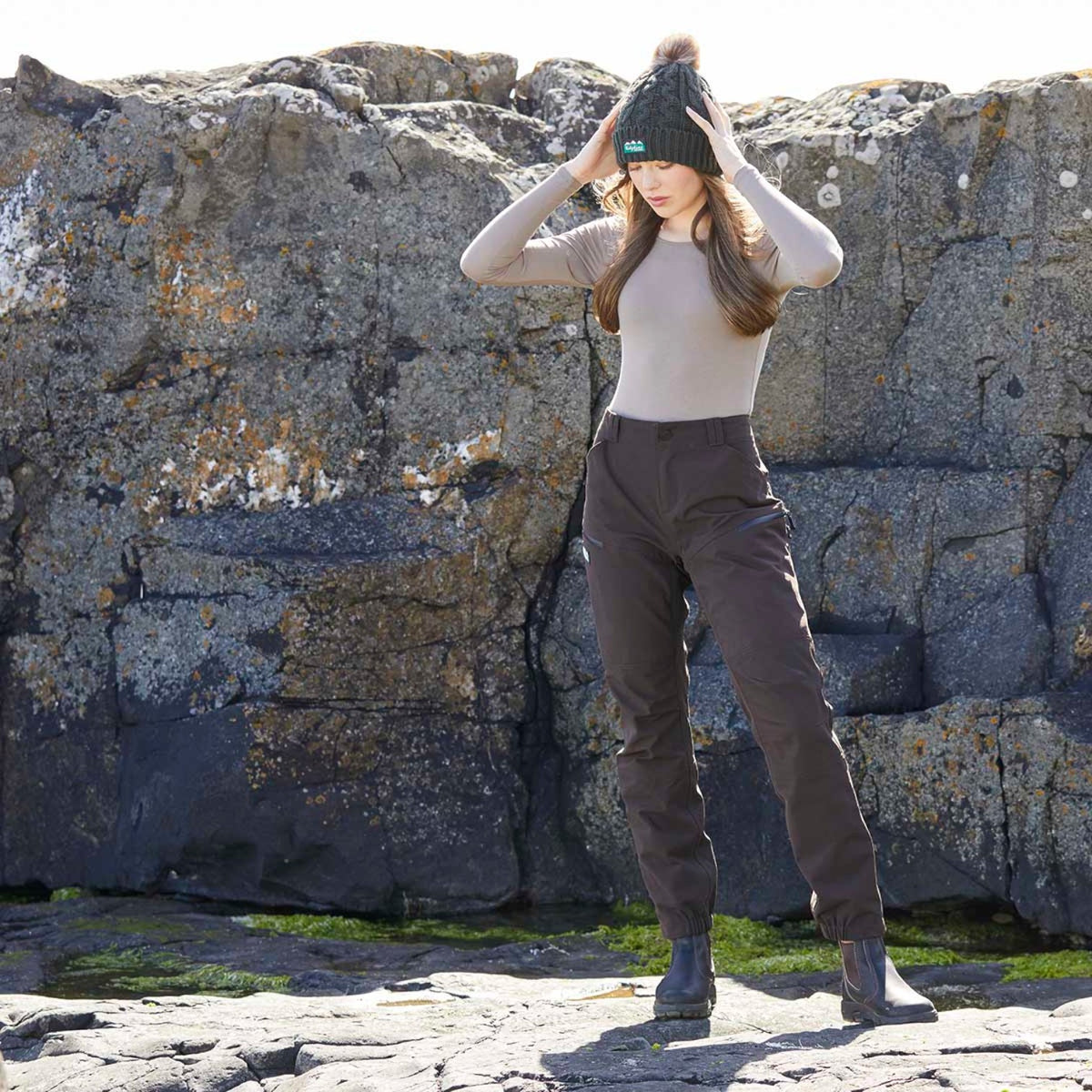 A woman stands outdoors against a large rock face, wearing a beige fitted long-sleeve top, dark brown pants, black boots, and a dark knit beanie with a pom-pom.