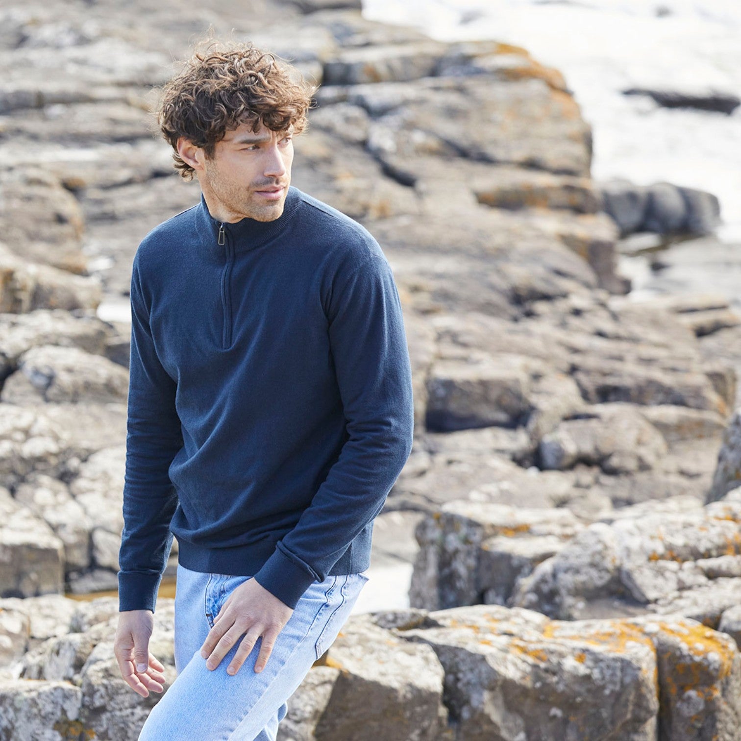 A man with curly brown hair wearing a navy blue quarter-zip long-sleeve sweater and light blue jeans, standing on a rocky coastal path.