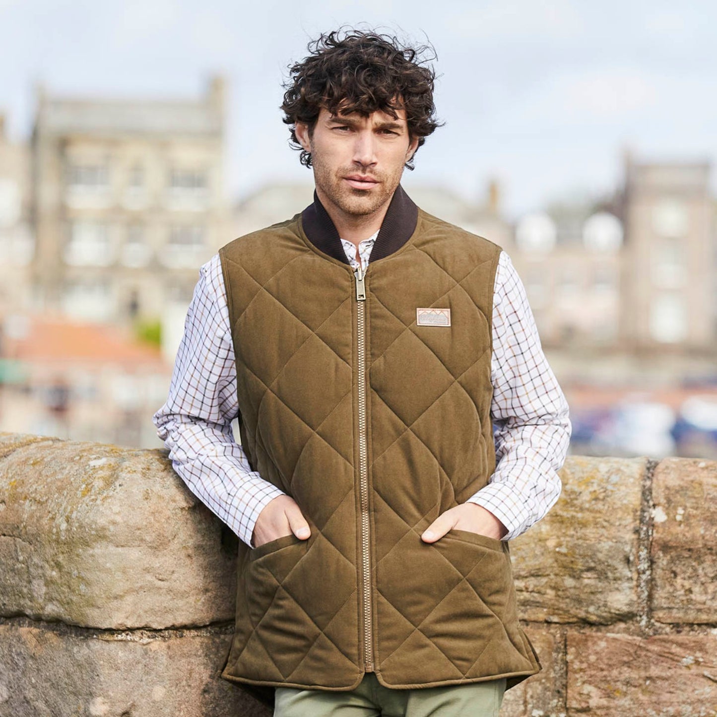 A man wearing a brown diamond-quilted zip-up vest over a white checkered shirt, leaning against a stone wall with a blurred city background.