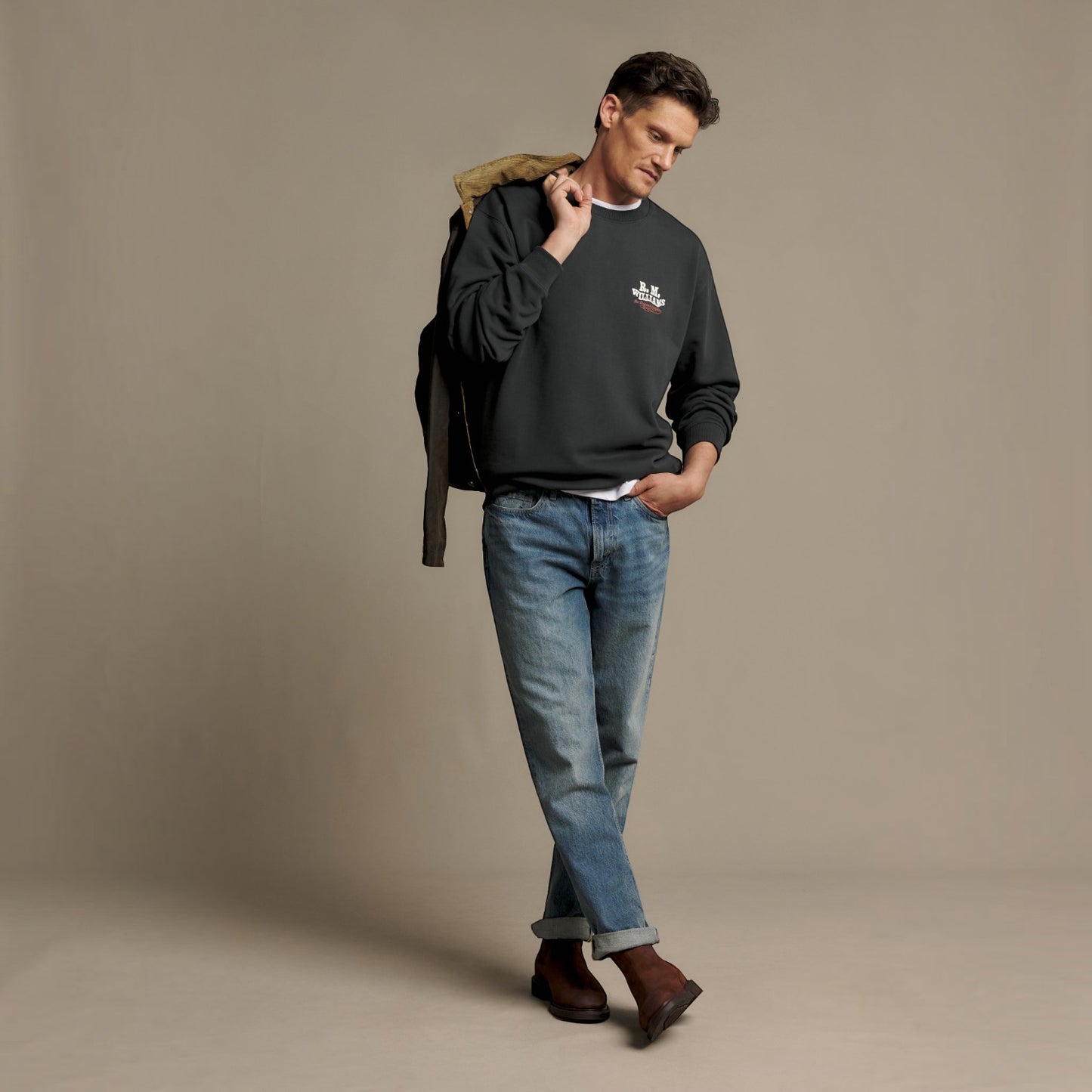 A man modeling a dark charcoal gray sweatshirt with a small white embroidered logo on the chest, styled with blue jeans and brown boots, standing against a solid beige studio background.