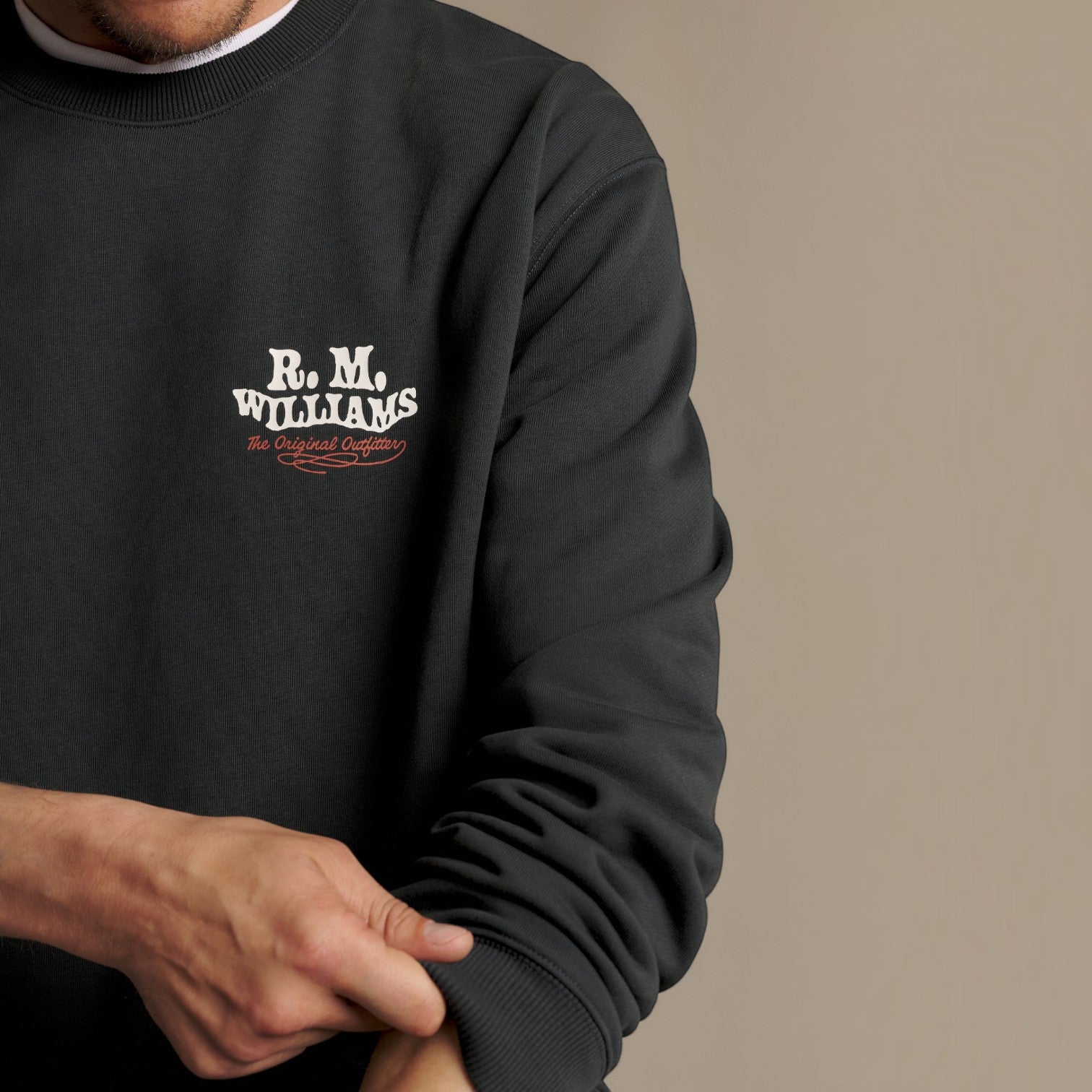 A close-up of a person wearing a dark charcoal crewneck sweatshirt with a white and red 'R.M.