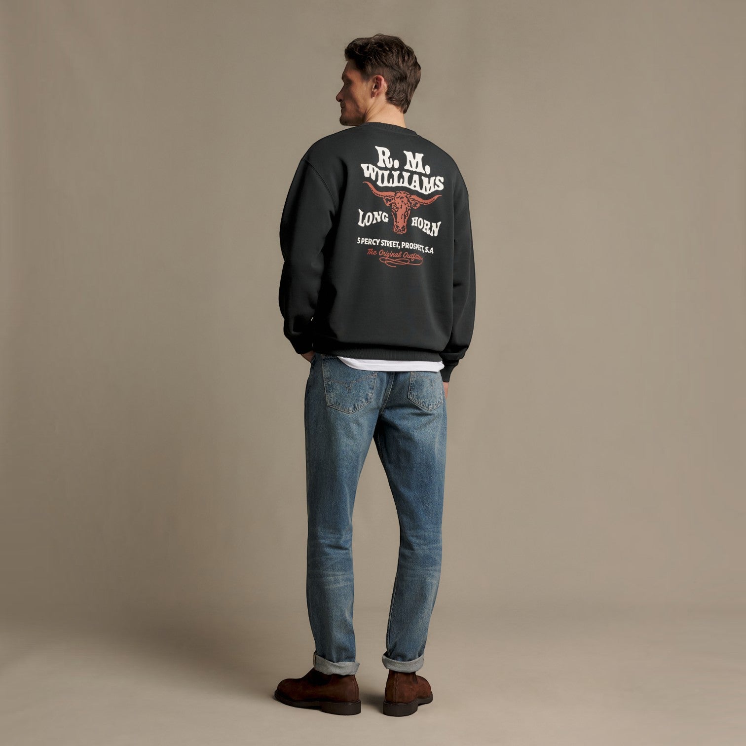 A man stands with his back to the camera wearing a black bomber jacket with a large white and red 'R.M.