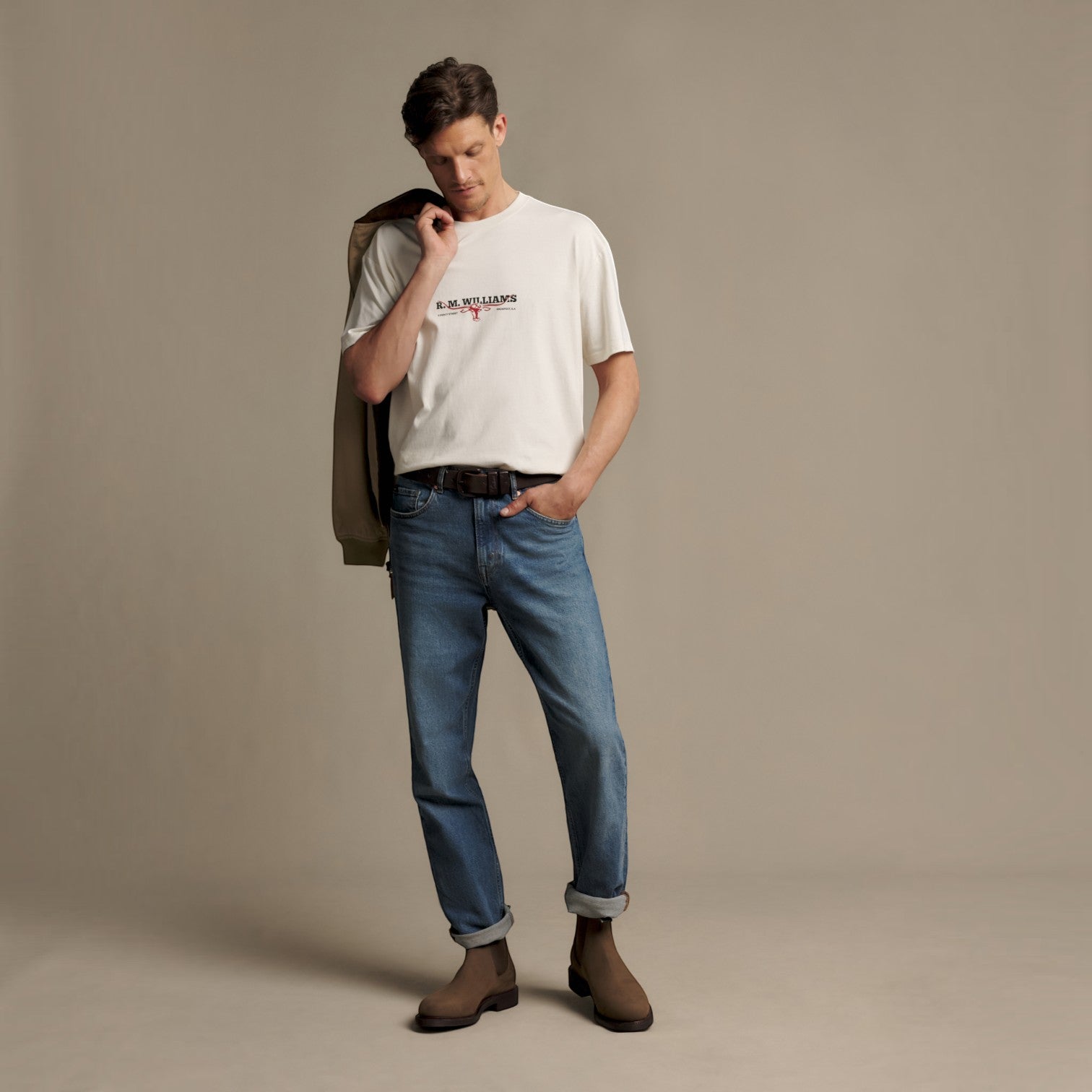 A man modeling a white graphic t-shirt with 'Burberry' printed on the chest, paired with blue jeans and brown boots, standing against a neutral beige studio backdrop.