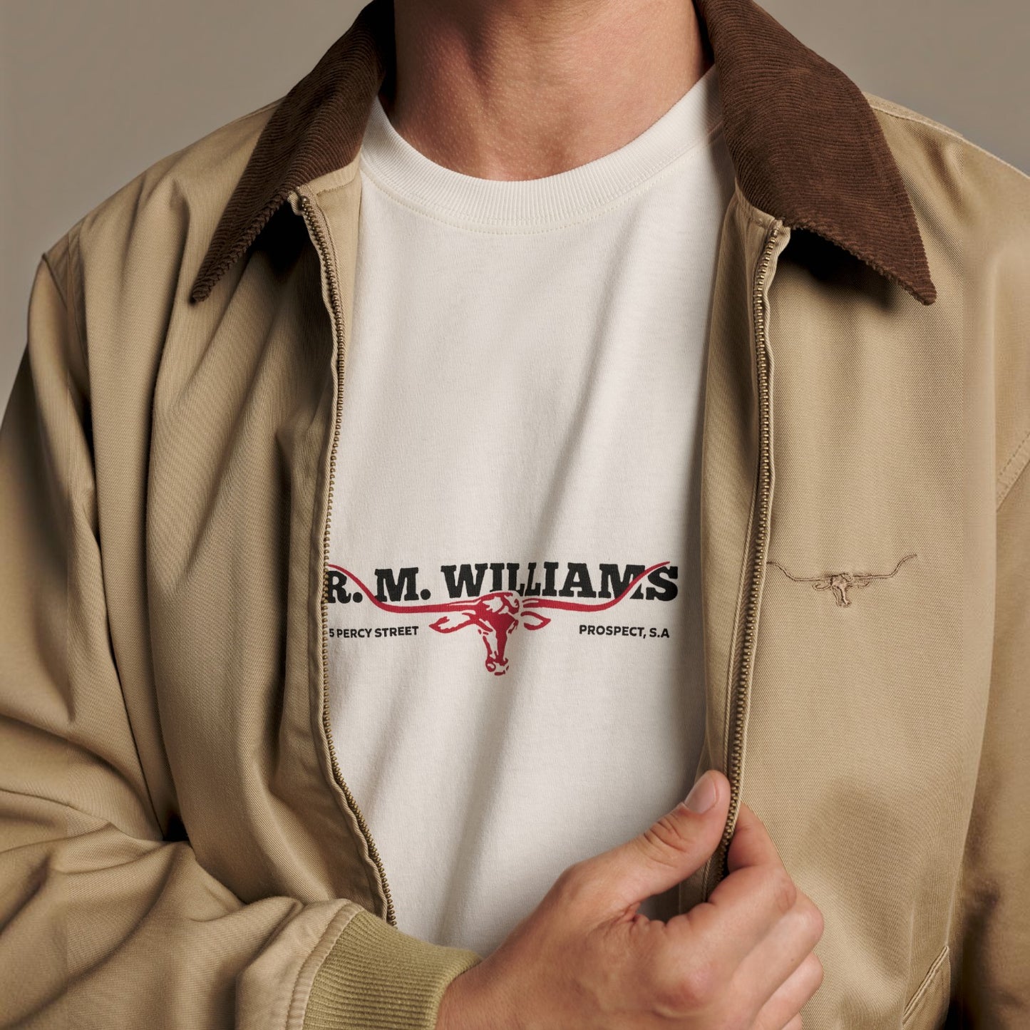 A close-up of a person wearing a beige zip-front jacket with a brown corduroy-style collar over a white graphic T-shirt. The shirt has black and red 'R.