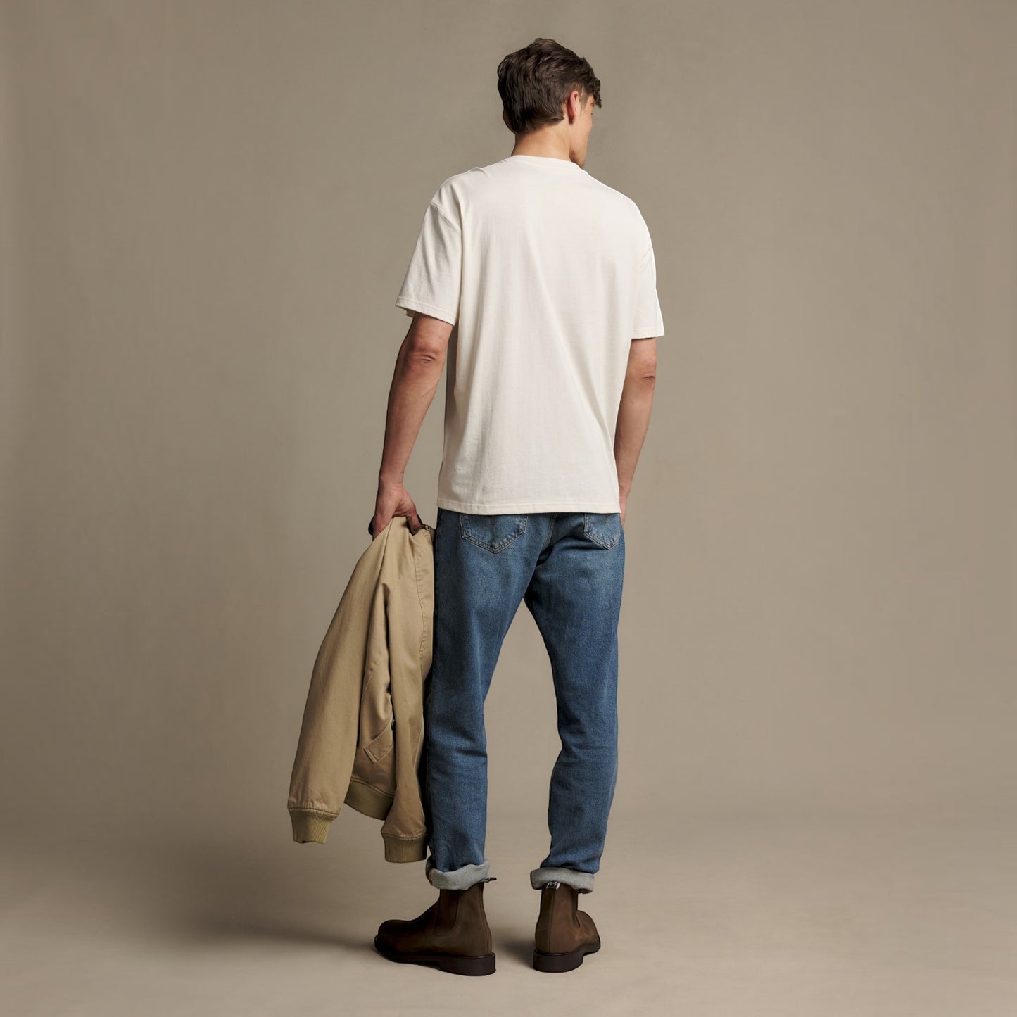A man stands with his back to the camera wearing a white crewneck t-shirt, blue jeans, and brown boots, holding a beige jacket in one hand against a plain beige studio background.