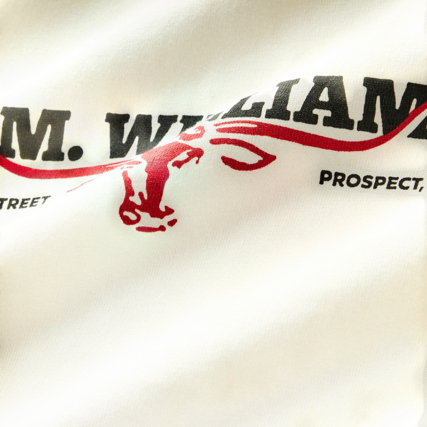 Close-up of a white fabric surface printed with bold black text reading "R.M. WILLIAMS" and a red graphic element, with partial black text reading "PROSPECT" and "STREET" nearby.