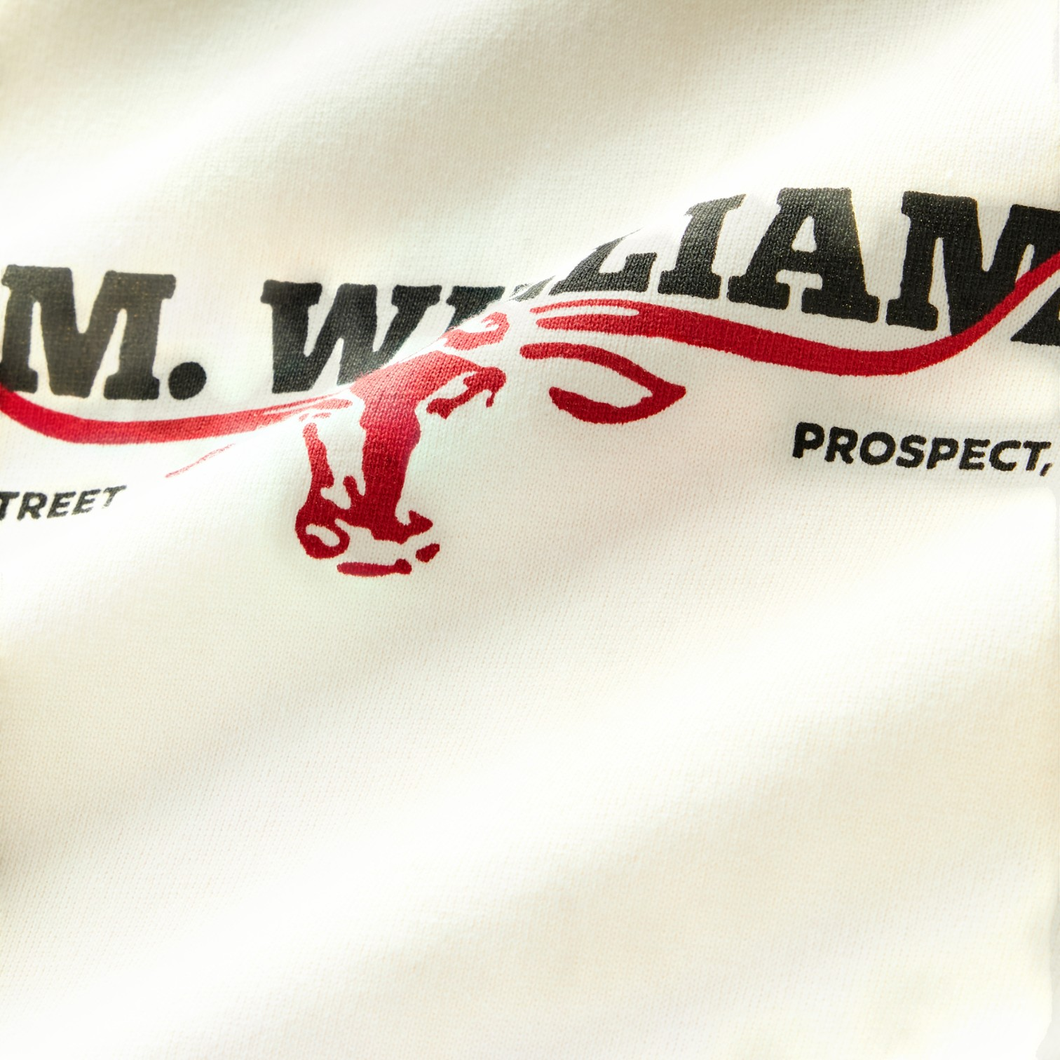 Close-up of a white fabric surface printed with bold black text reading "R.M. WILLIAMS" and a red graphic element, with partial black text reading "PROSPECT" and "STREET" nearby.