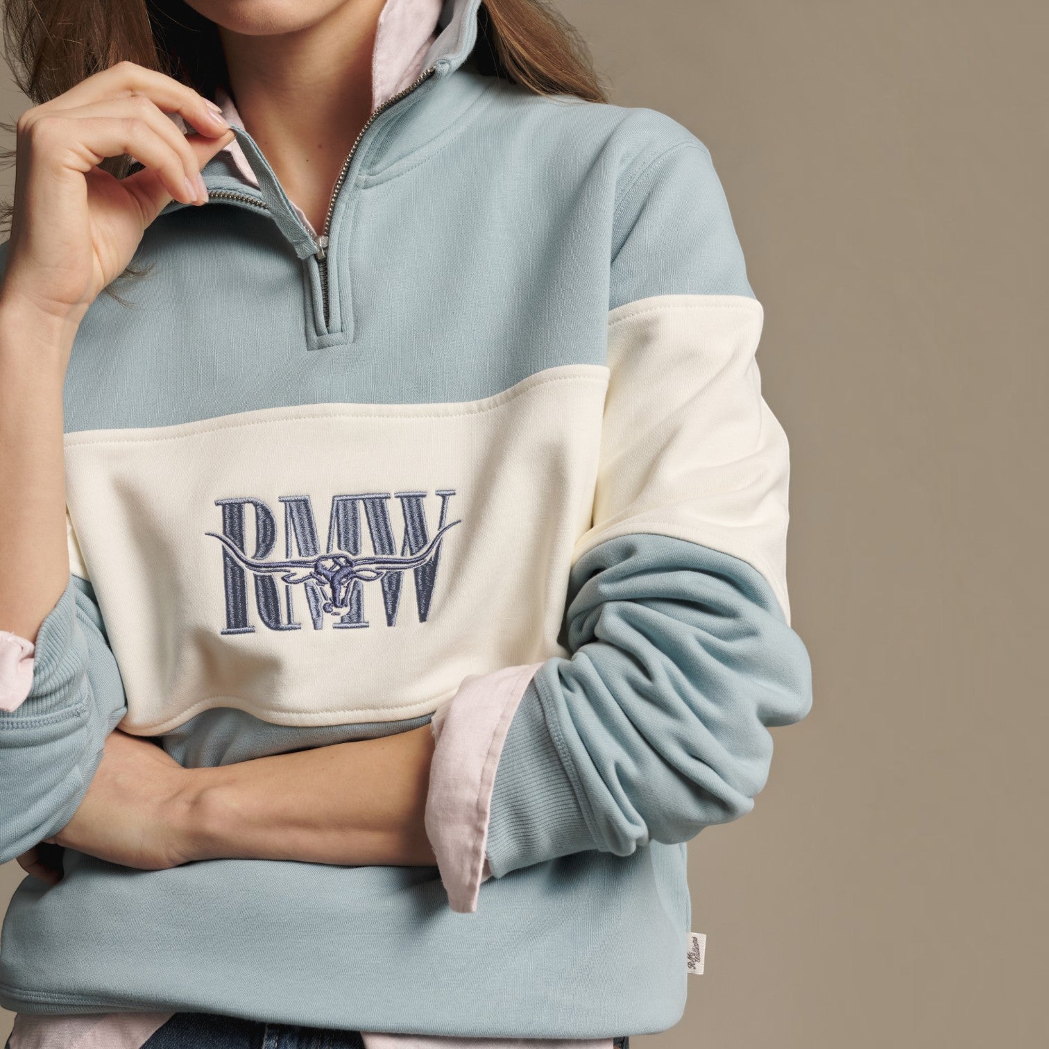 A close-up of a person wearing a color-blocked quarter-zip sweatshirt. The sweatshirt features a light blue body, an off-white horizontal chest stripe, and navy blue 'RAW' logo embroidery with a steer skull graphic on the chest.
