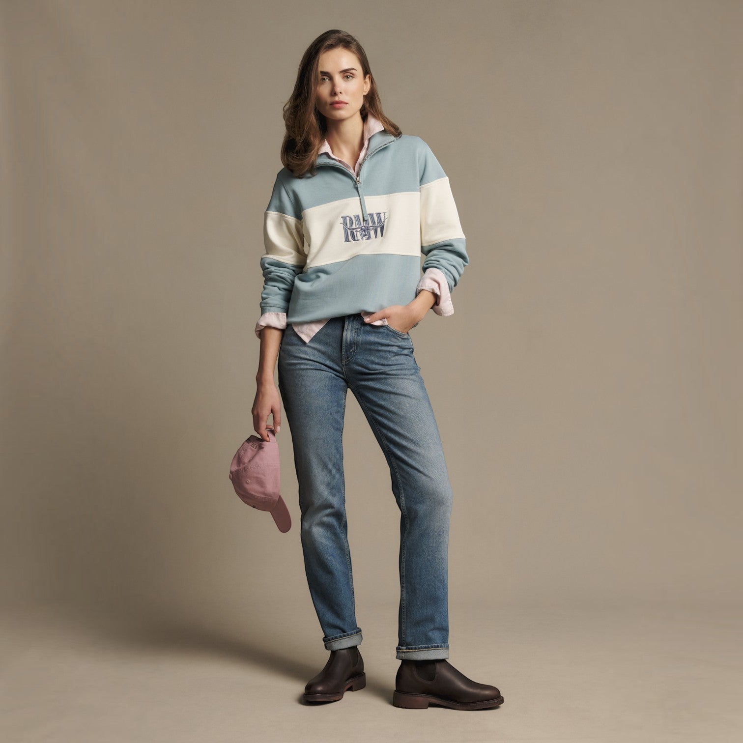 A woman stands in a studio wearing a color-blocked quarter-zip pullover with 'DARKLY DESIRABLE' printed across the chest, blue jeans, dark brown boots, and holding a dusty pink cap. The background is a solid warm beige studio backdrop.