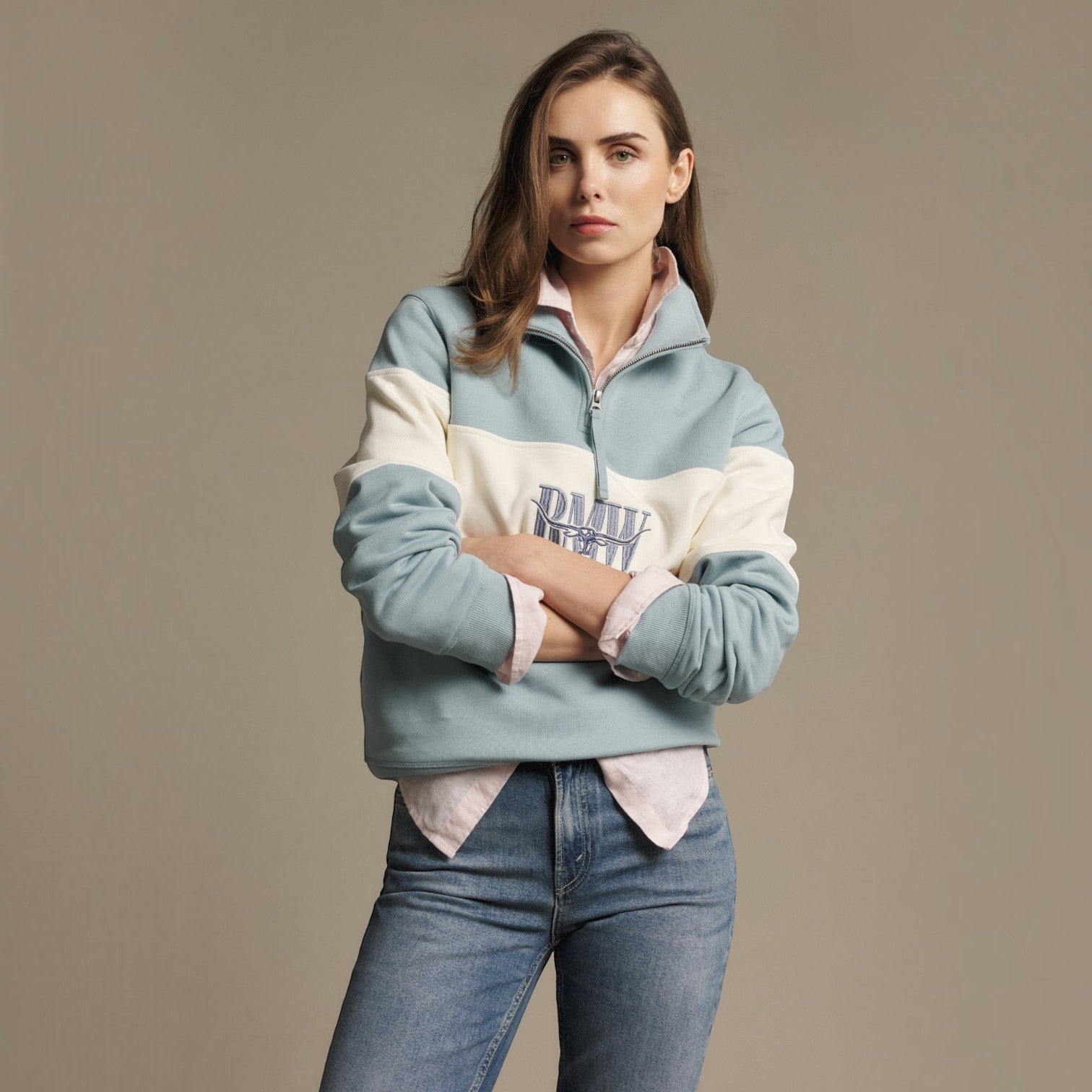 A woman modeling a color-blocked quarter-zip sweatshirt in light blue, cream, and white, layered over a light pink shirt and paired with blue jeans, against a solid taupe background.