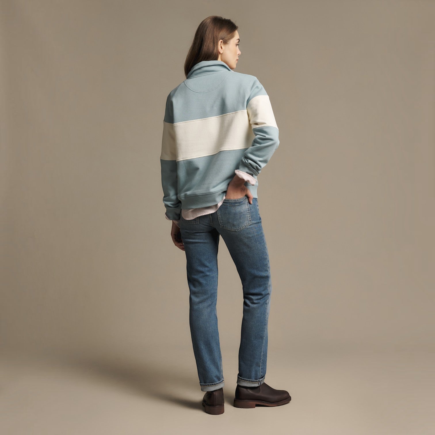 A model shown from the back wearing a light blue and cream color-blocked fleece jacket, blue jeans, and dark brown boots against a solid beige studio background.