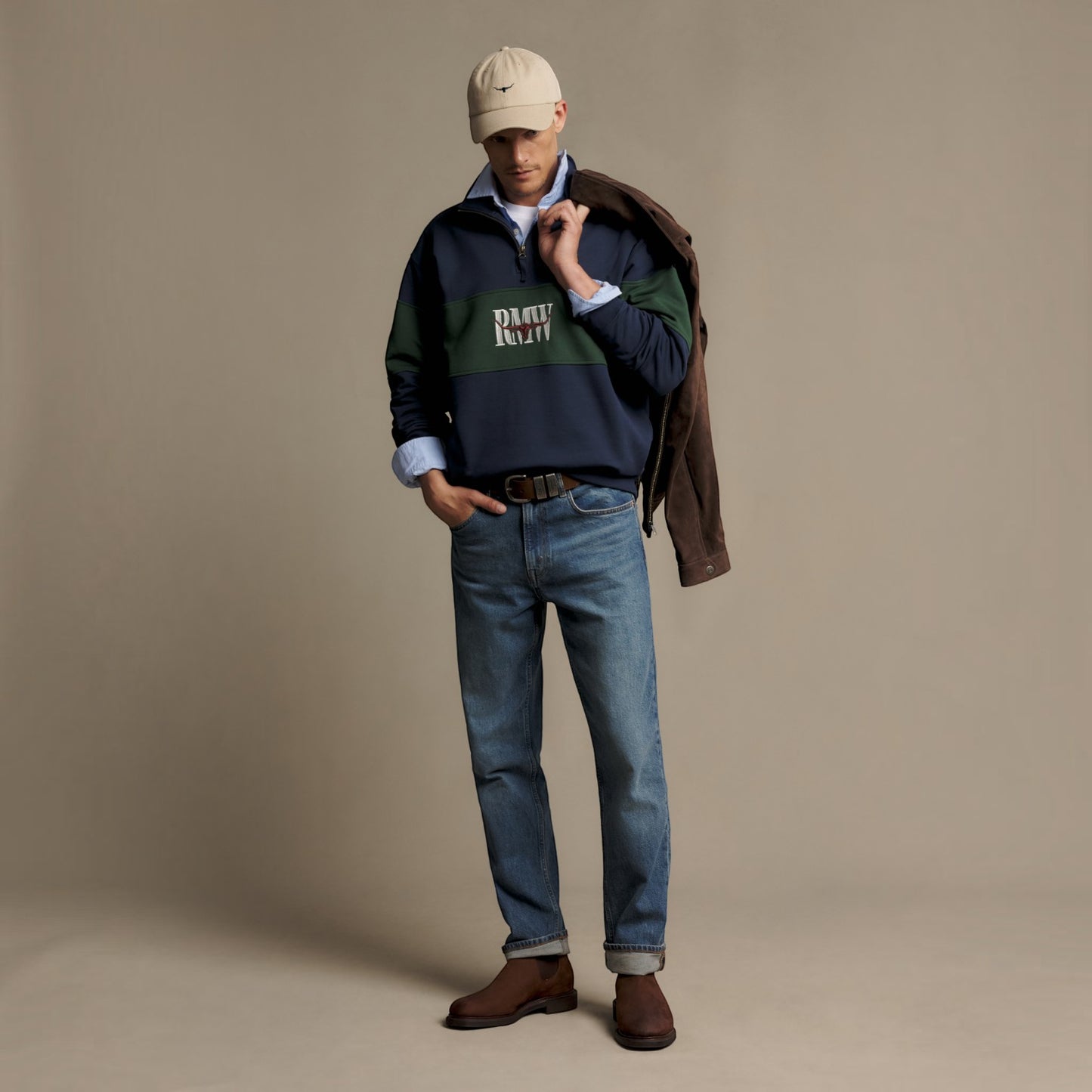 A male model stands in a studio wearing a navy and green color-blocked sweatshirt with a 'DAILY RUTT' logo, blue jeans, brown leather boots, a beige baseball cap, and a brown jacket draped over his shoulder, against a solid beige backdrop.