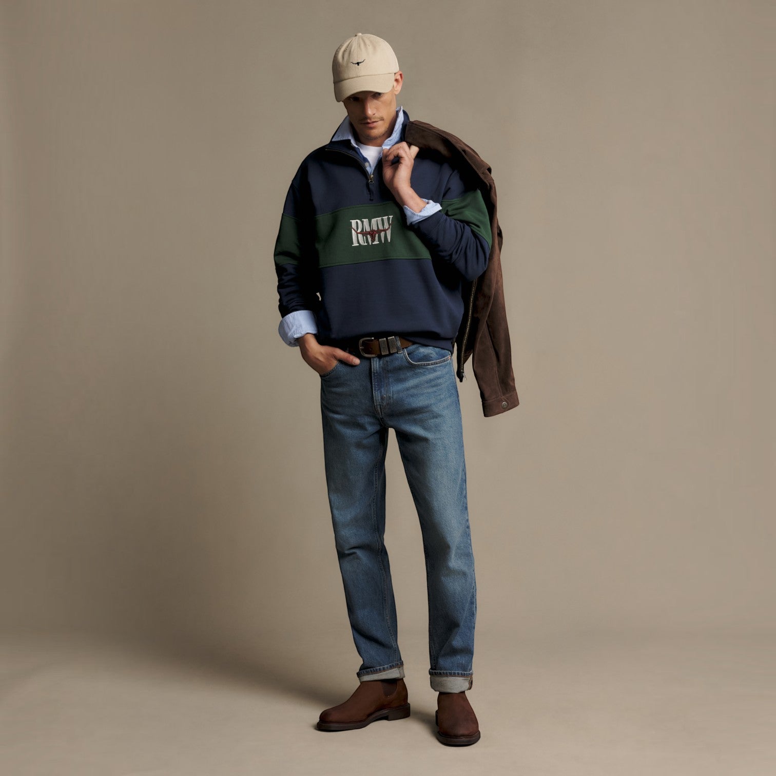 A male model stands in a studio wearing a navy and green color-blocked sweatshirt with a 'DAILY RUTT' logo, blue jeans, brown leather boots, a beige baseball cap, and a brown jacket draped over his shoulder, against a solid beige backdrop.