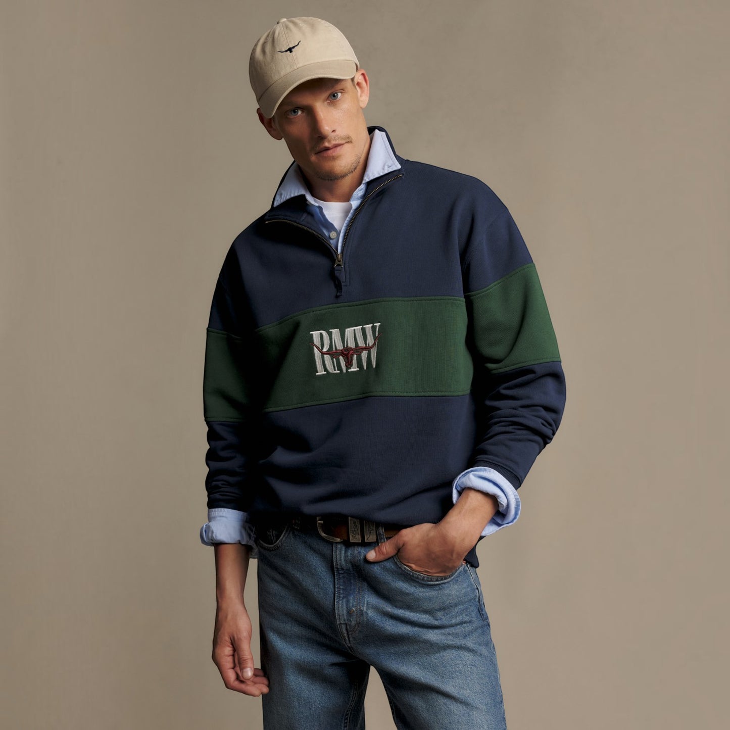 A man wearing a navy and green color-blocked quarter-zip sweatshirt with 'RMW' embroidered on the chest, styled with blue jeans, a light blue collared shirt, and a beige baseball cap against a solid beige background.