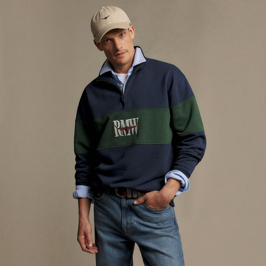 A man wearing a navy and green color-blocked quarter-zip sweatshirt with 'RMW' embroidered on the chest, styled with blue jeans, a light blue collared shirt, and a beige baseball cap against a solid beige background.