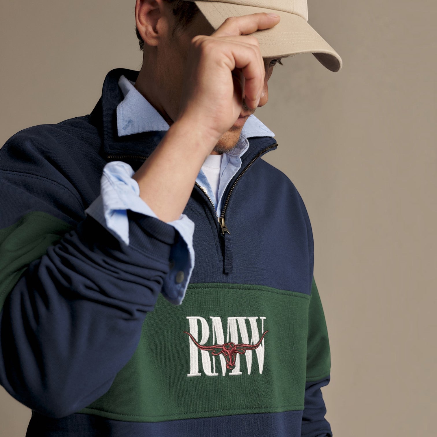 A close-up of a person wearing a navy and green color-block quarter-zip pullover with a white and red 'RMW' bull logo on the chest, layered over a light blue collared shirt and a beige cap.