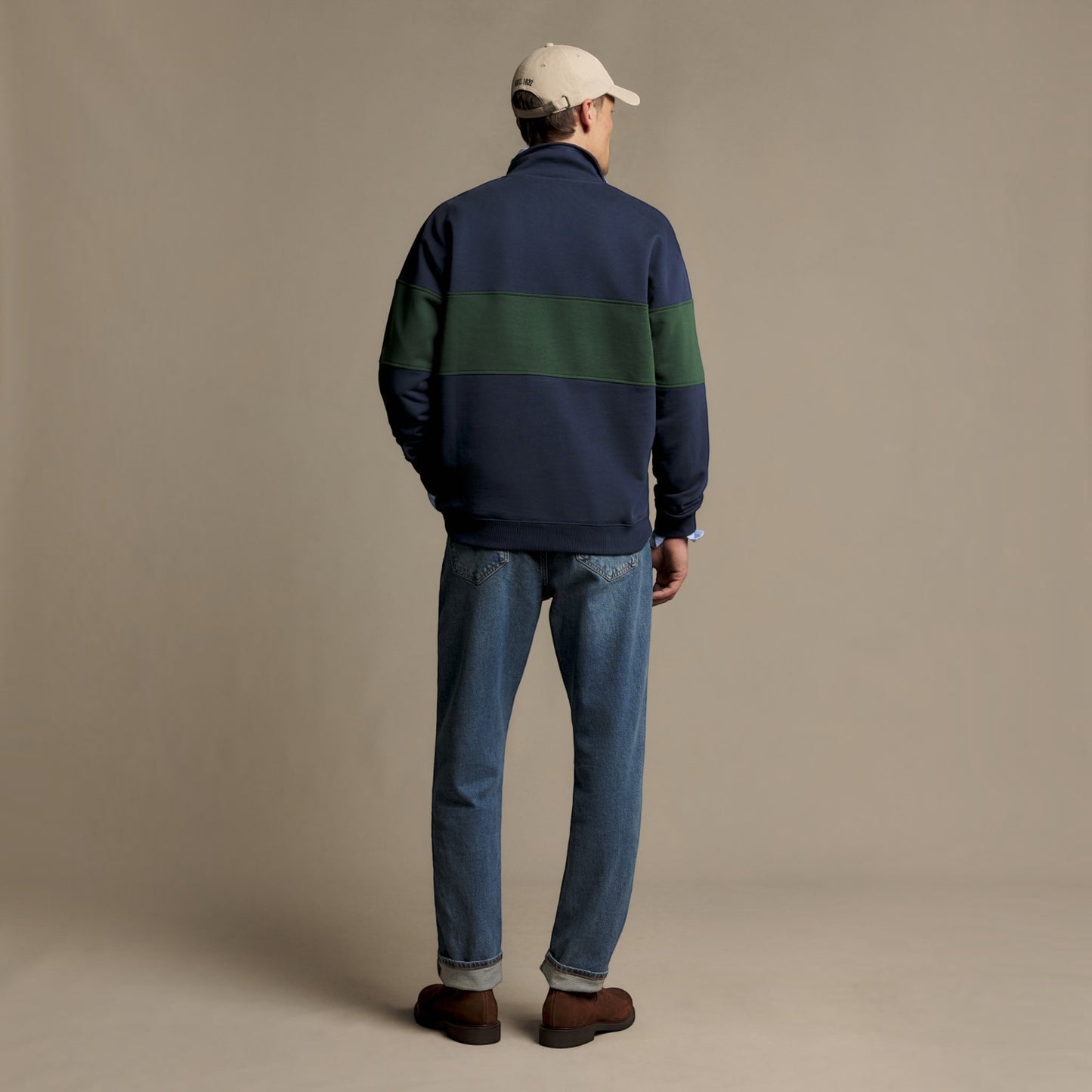 A man viewed from behind wearing a navy blue quarter-zip sweatshirt with a dark green horizontal stripe across the upper back, blue jeans, brown shoes, and a beige baseball cap, standing against a solid beige studio background.