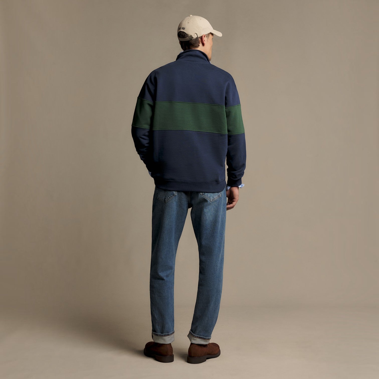 A man viewed from behind wearing a navy blue quarter-zip sweatshirt with a dark green horizontal stripe across the upper back, blue jeans, brown shoes, and a beige baseball cap, standing against a solid beige studio background.