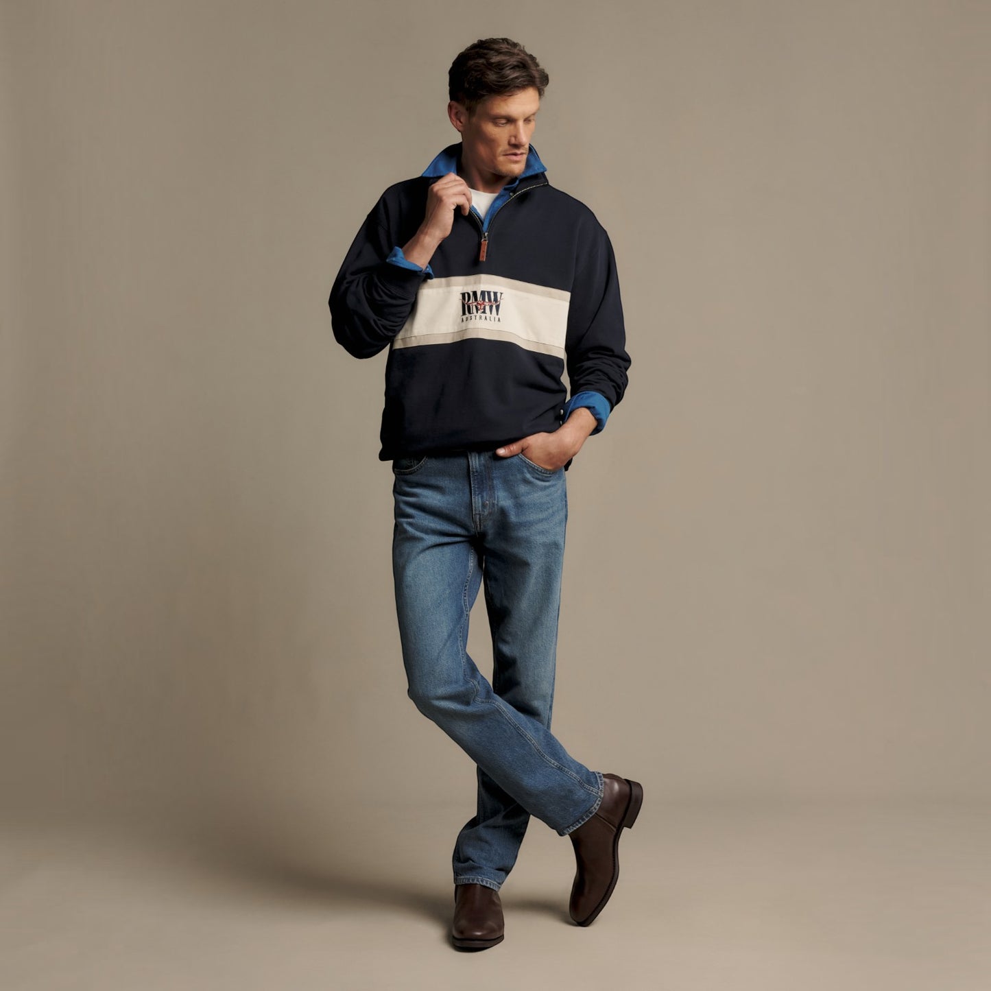 A man modeling a navy blue quarter-zip pullover sweatshirt with a wide cream-colored chest stripe and 'RMW' printed on it, paired with blue jeans and brown boots, against a solid beige studio background.