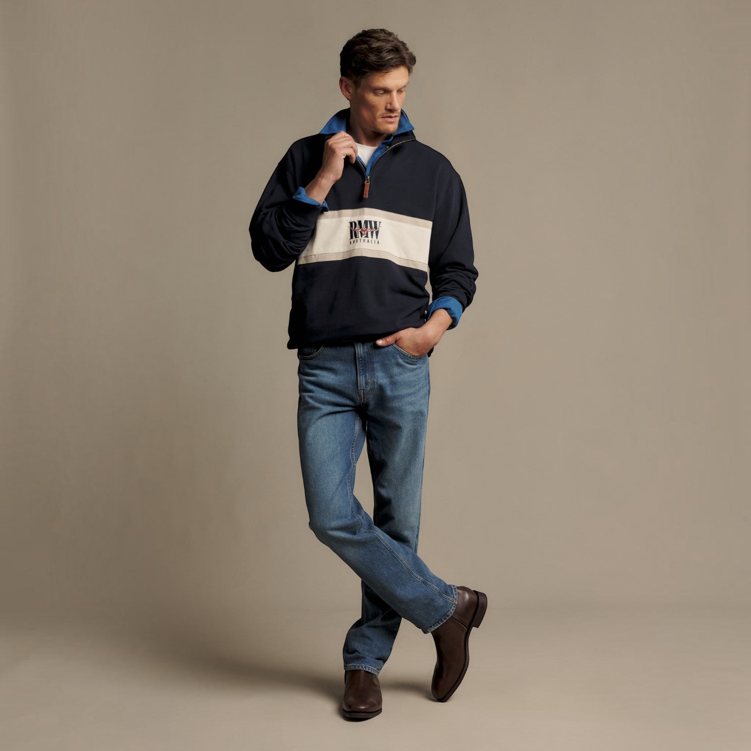 A man modeling a navy blue quarter-zip pullover sweatshirt with a wide cream-colored chest stripe and 'RMW' printed on it, paired with blue jeans and brown boots, against a solid beige studio background.