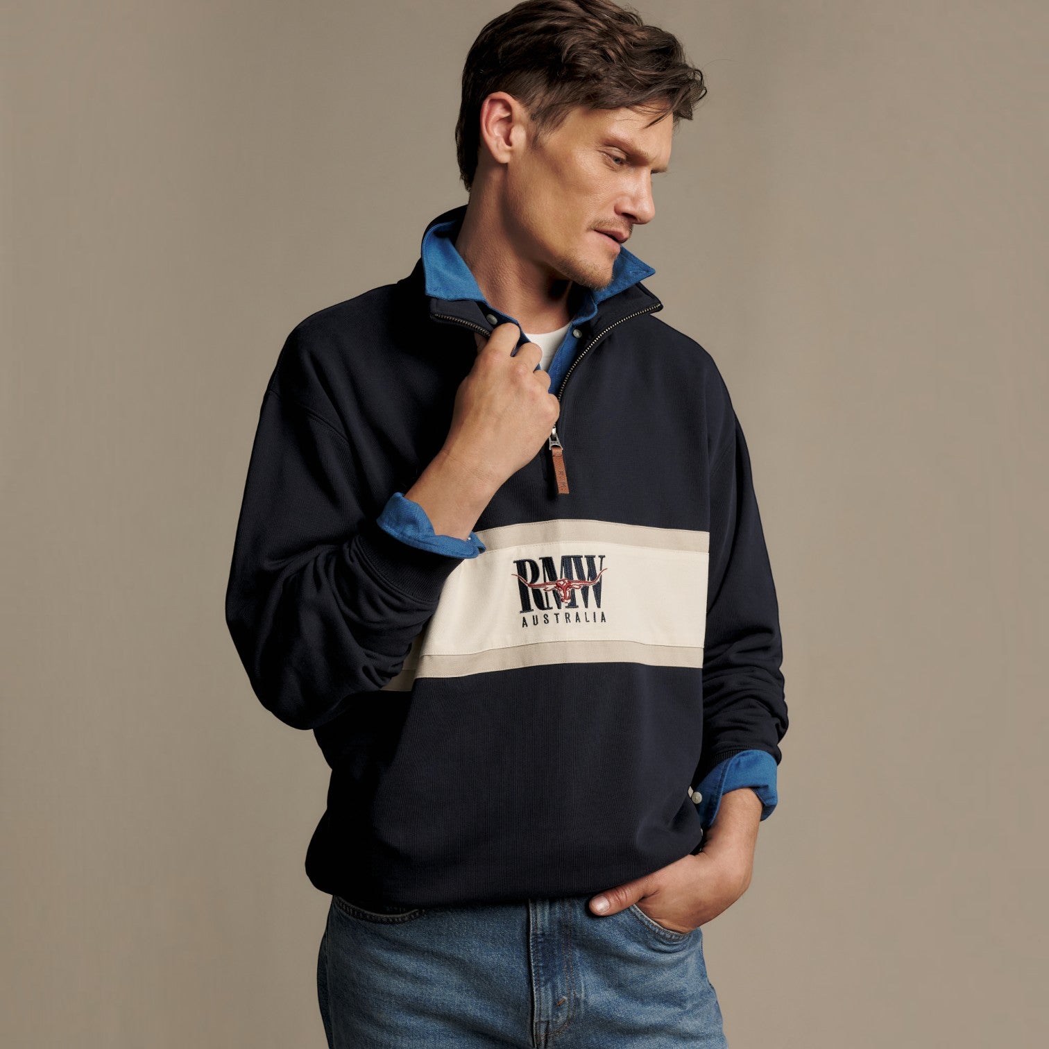 A man wearing a navy blue quarter-zip sweatshirt with a wide beige chest panel and blue inner collar lining, featuring a RMW Australia logo, paired with blue jeans against a neutral studio background.