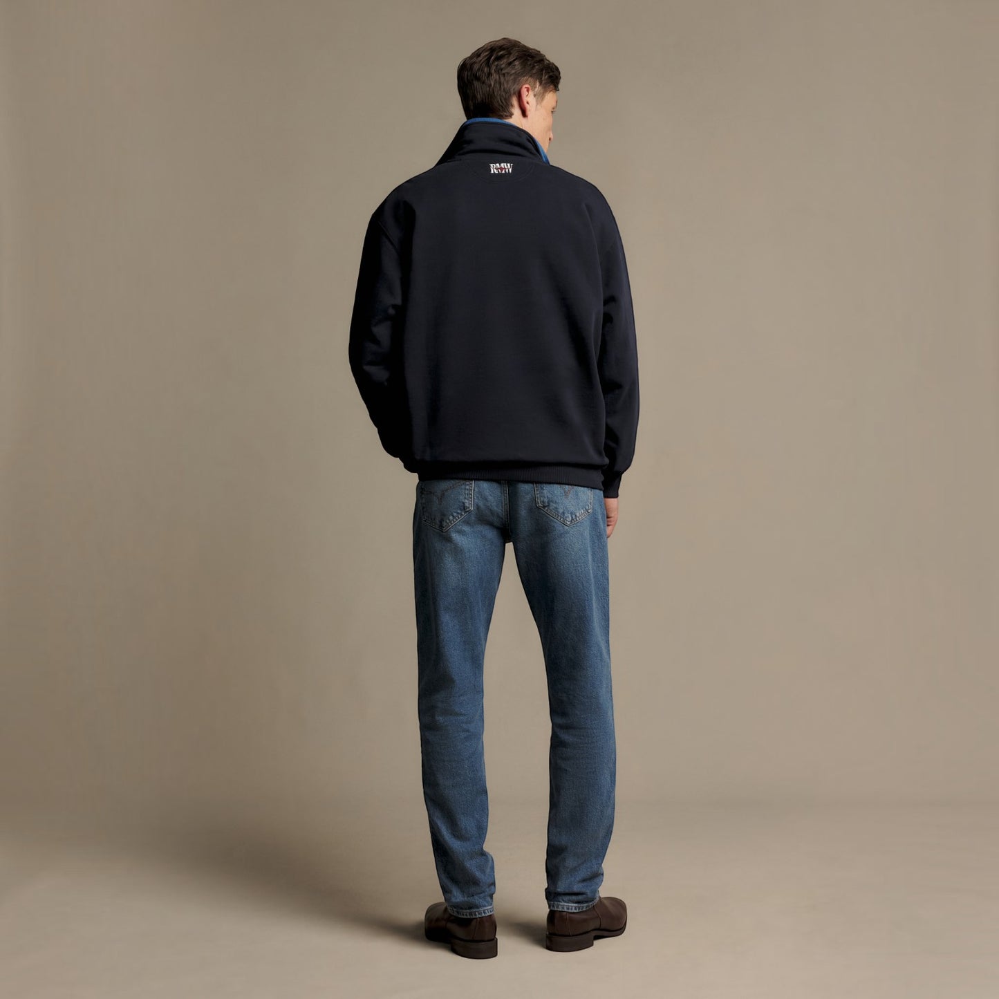 A man viewed from behind wearing a dark navy quarter-zip fleece jacket, blue jeans, and brown shoes, standing against a solid beige studio background.