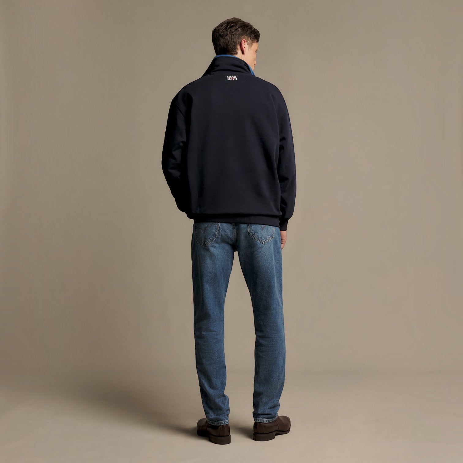 A man viewed from behind wearing a dark navy quarter-zip fleece jacket, blue jeans, and brown shoes, standing against a solid beige studio background.