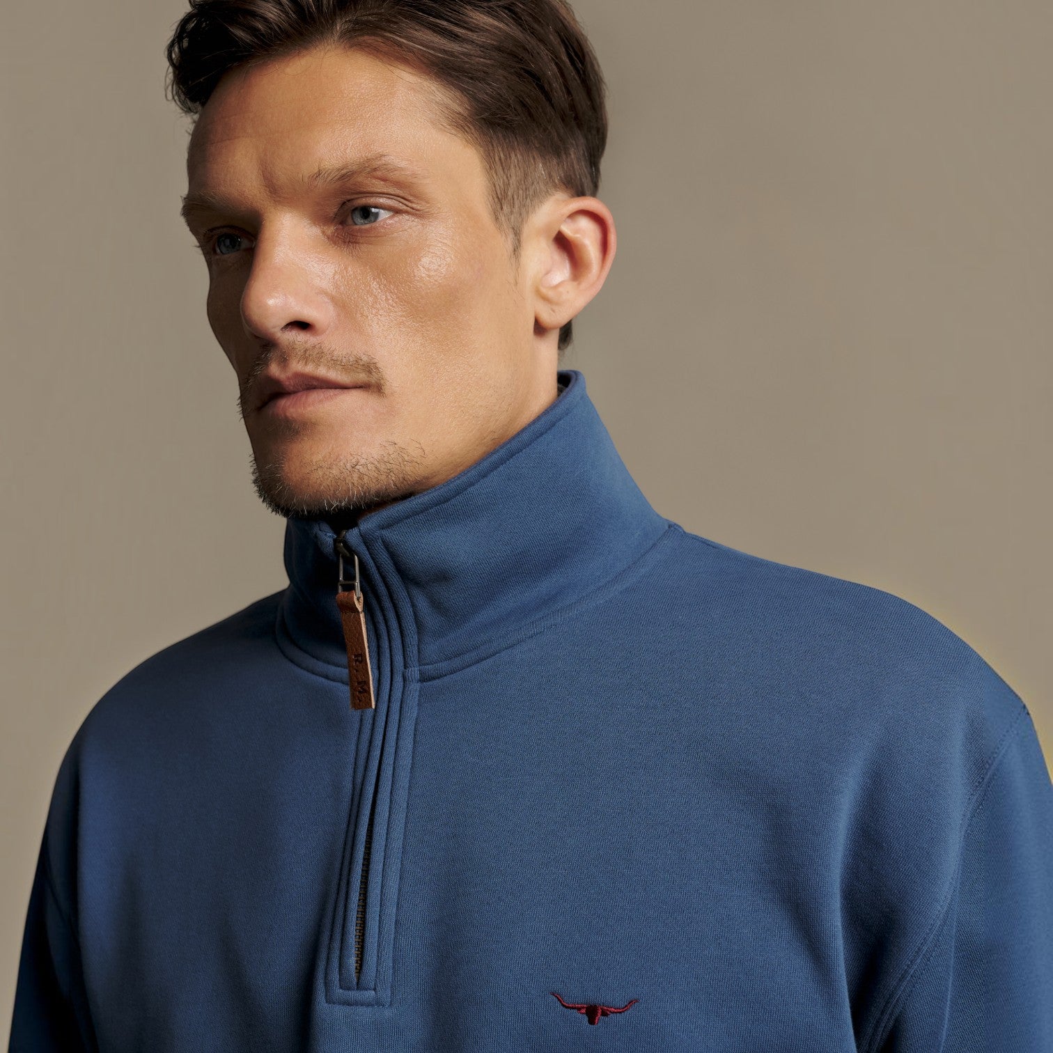 A close-up of a man wearing a blue quarter-zip mock neck sweater with a small embroidered logo on the chest, set against a solid beige background.