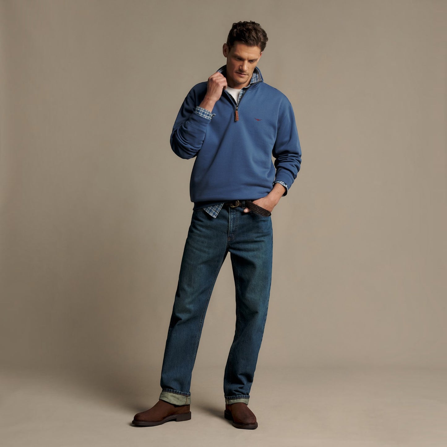 A man modeling a blue quarter-zip pullover sweater with a plaid collar, paired with blue jeans and brown leather shoes, standing against a solid beige studio background.