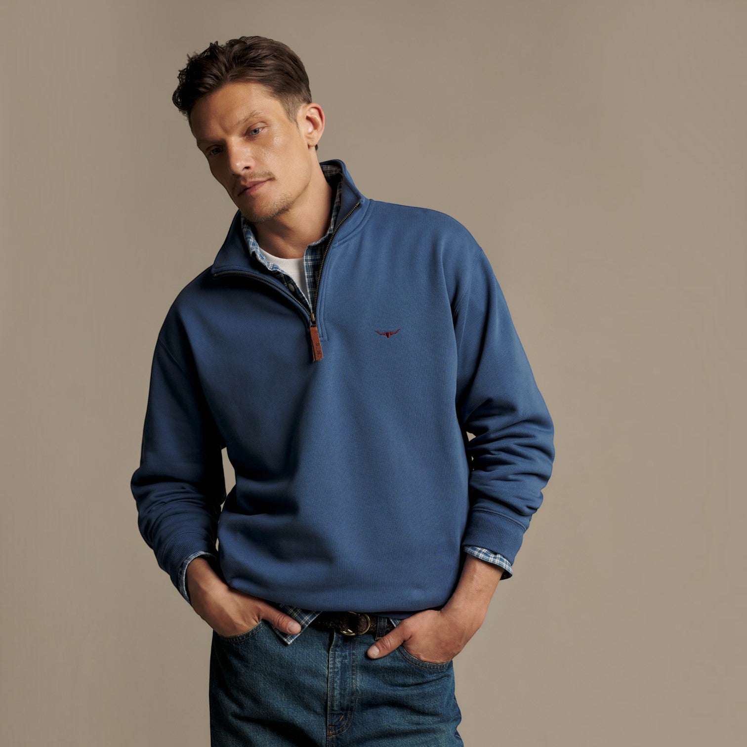 A man wearing a blue quarter-zip pullover sweater with a small embroidered bird logo on the chest, styled over a plaid shirt and jeans, against a solid beige studio background.